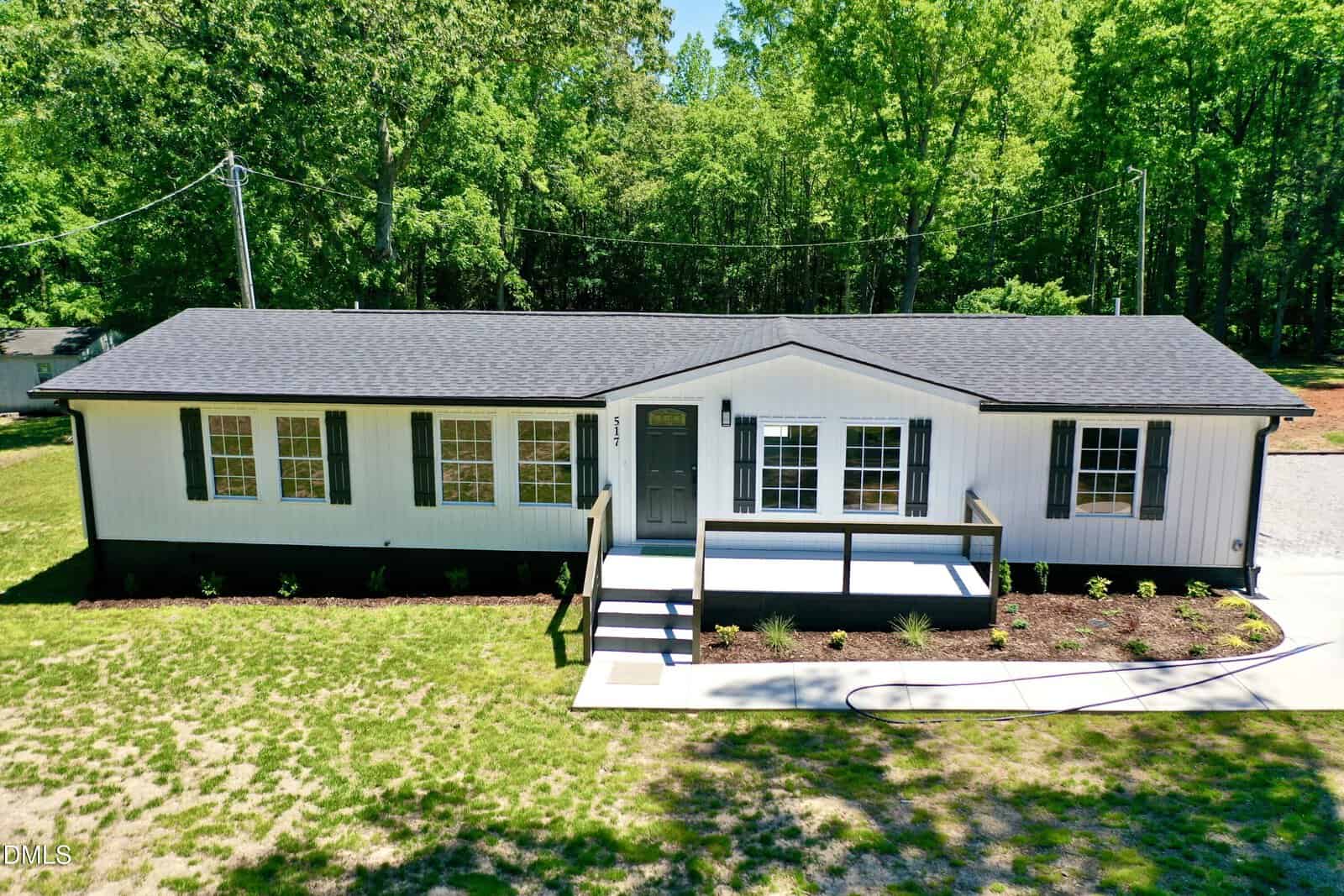 A white, single-story manufactured home with black shutters and a front porch sits on a grassy yard, surrounded by trees and a paved driveway under bright sunlight.