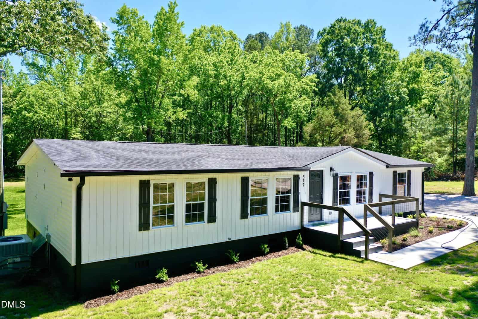 A single-story white house with black trim, a ramp leading to the front door, and large windows, set against a backdrop of tall green trees and a well-kept lawn.