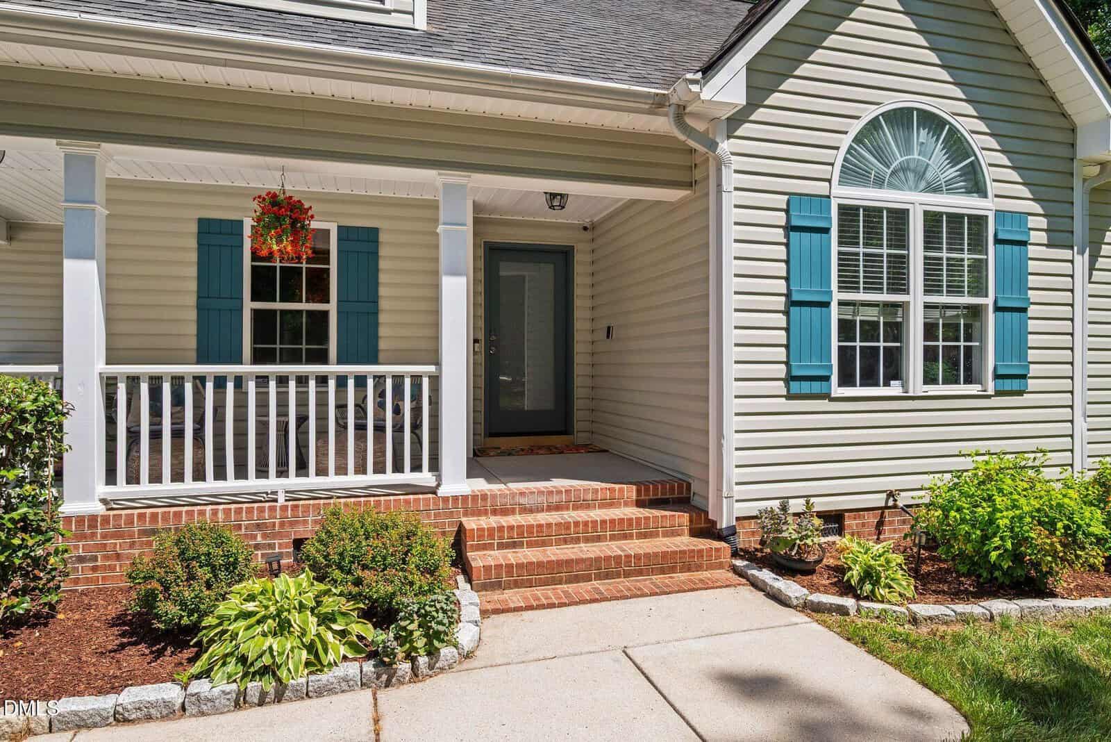 A front porch with brick steps and white railing leads to a dark front door. Blue shutters frame a large arched window. Landscaping with green shrubs and a hanging flower basket add charm.