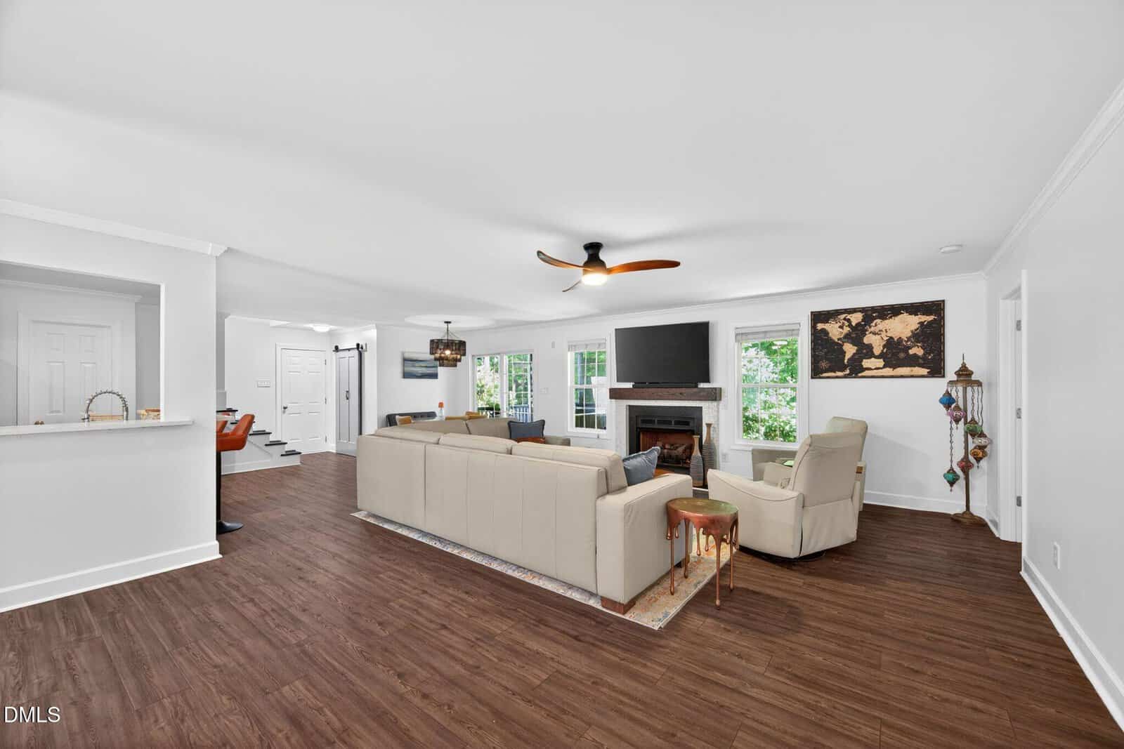 Modern living room with a beige sectional sofa, ceiling fan, fireplace with TV above, large windows, wooden floors, and a map wall art. The space is open to the entryway and kitchen with white walls throughout.