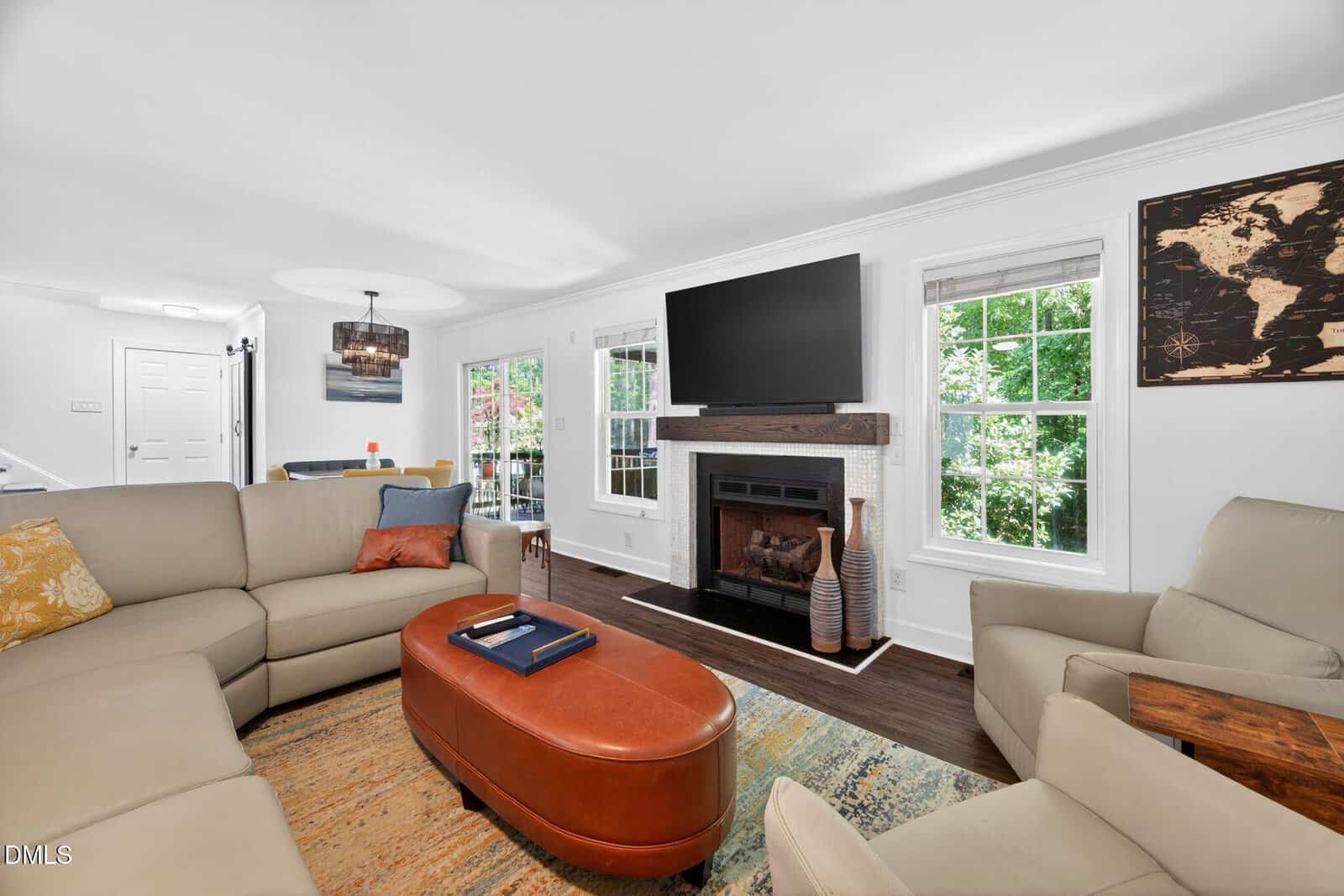 A bright living room with beige sofas, an orange ottoman, a fireplace with a TV above it, large windows with green outdoor views, hardwood floors, and a map on the wall. Dining area visible in the background.