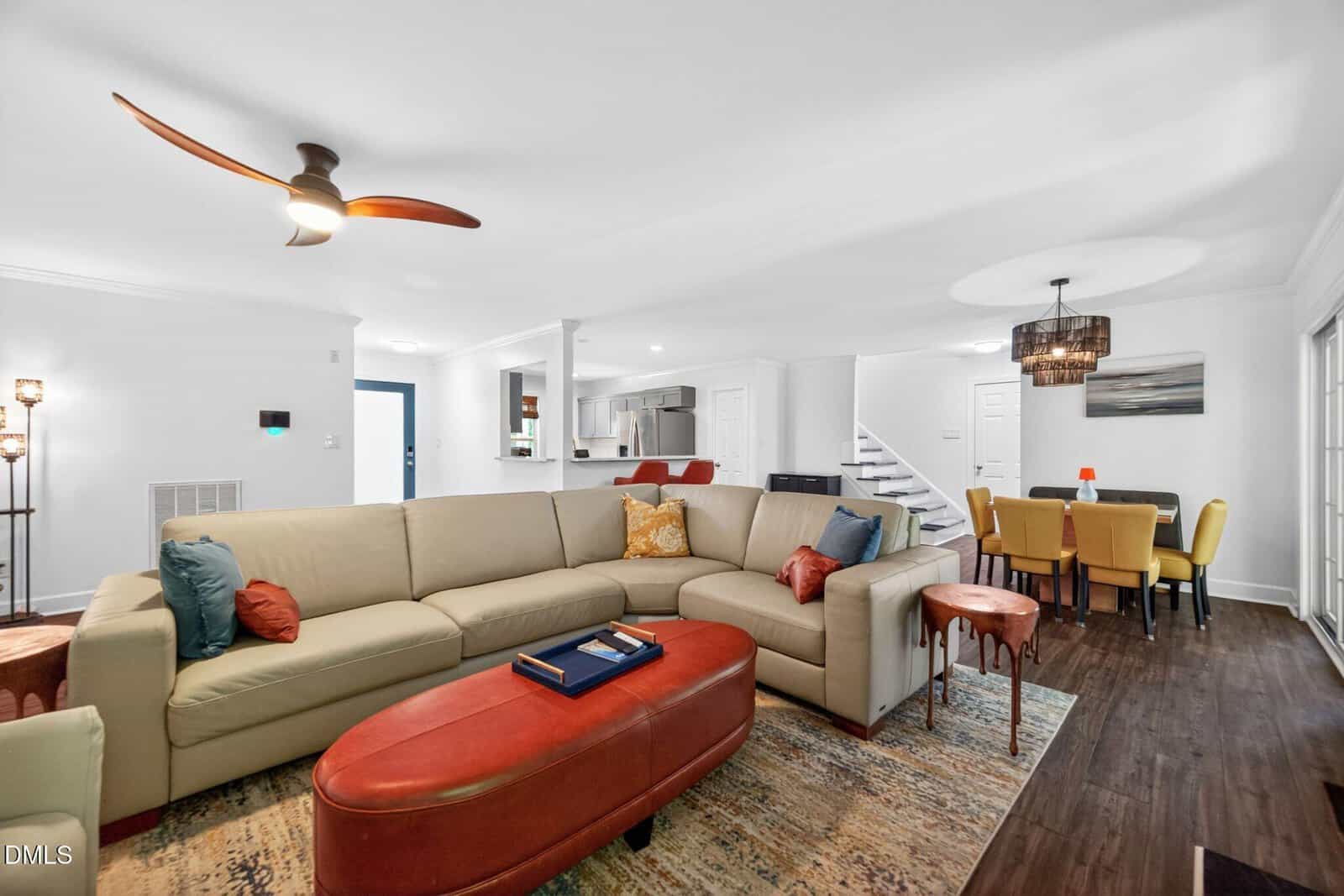 A spacious living and dining area with a large beige sectional sofa, colorful pillows, a red ottoman, modern ceiling fan, dining table with yellow chairs, and a staircase leading upstairs. The decor is contemporary and inviting.