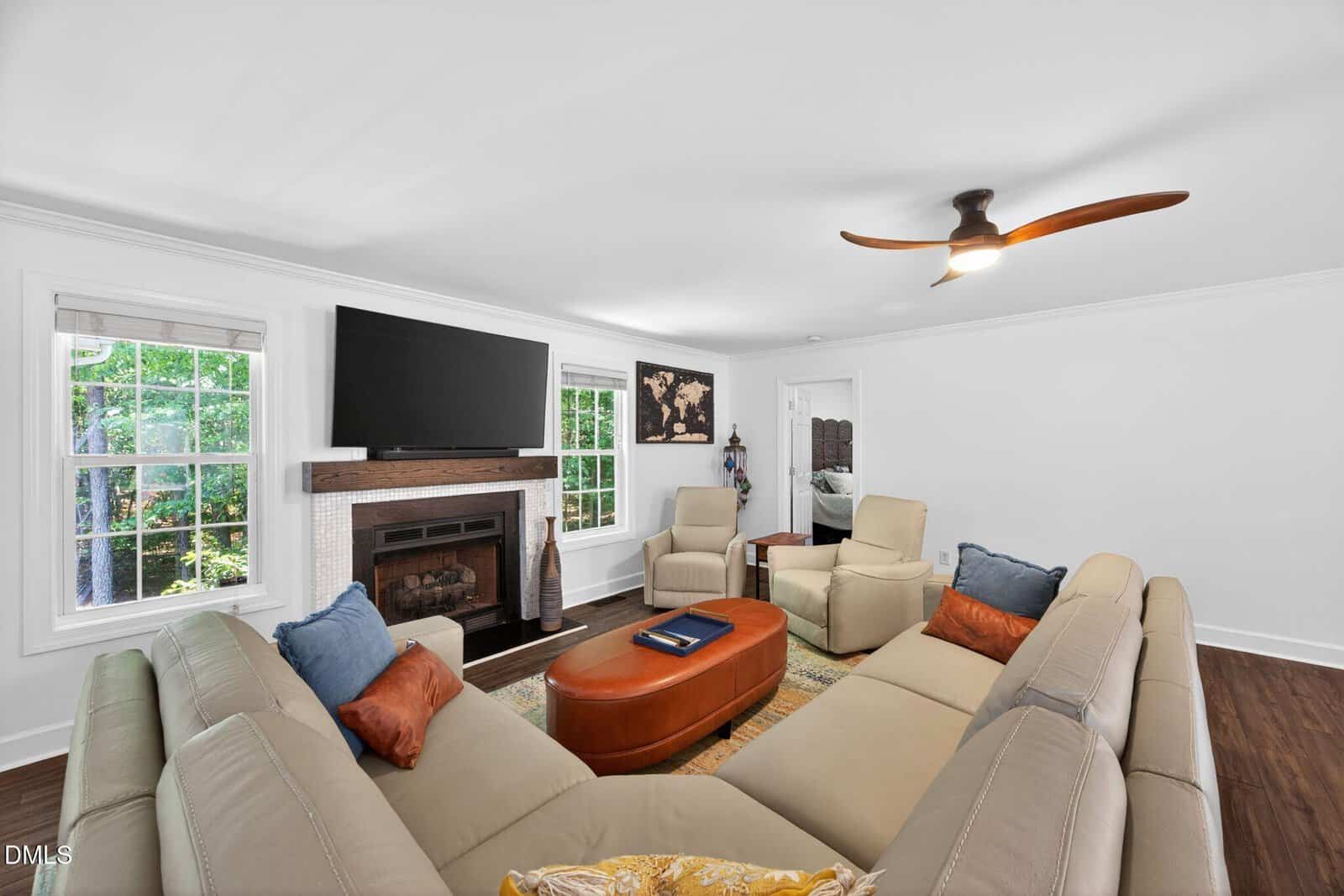 Bright living room with a large beige sectional sofa, colorful pillows, a brown ottoman, TV above a fireplace, windows showing greenery, and a ceiling fan with wooden blades. Artwork and decorative items are on the walls.
