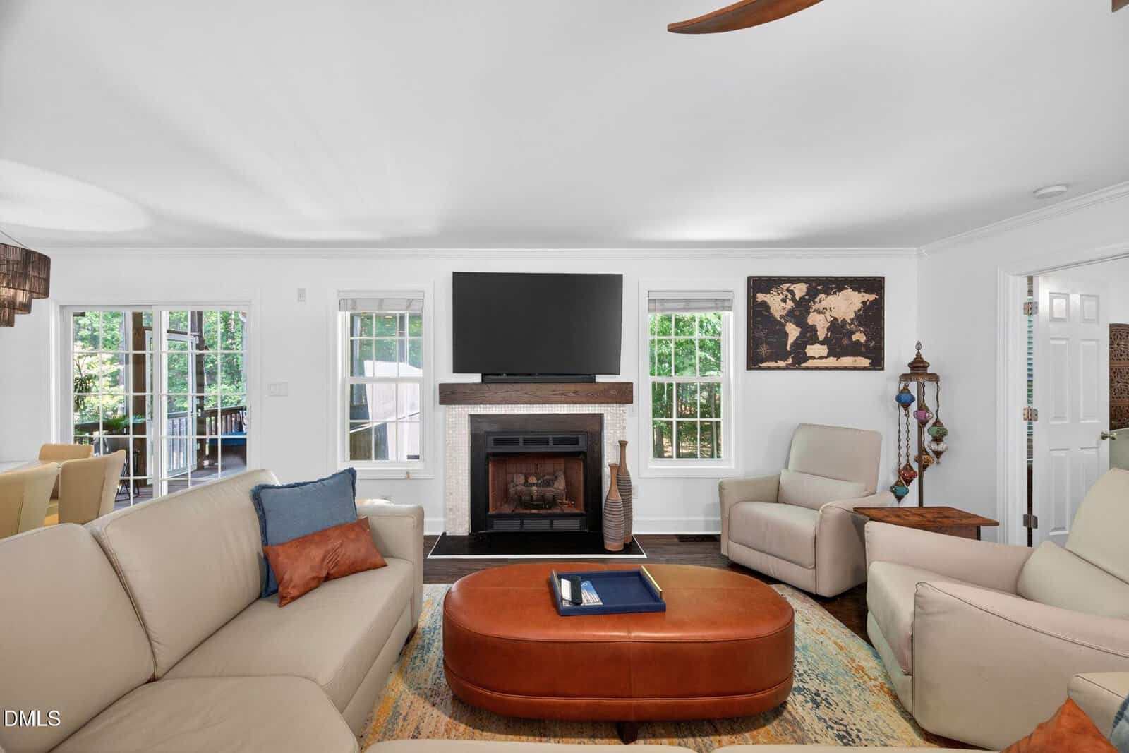 Bright living room with beige sofas, a brown ottoman, a fireplace under a wall-mounted TV, a world map on the wall, and large windows letting in natural light. Door opens to an outdoor area.