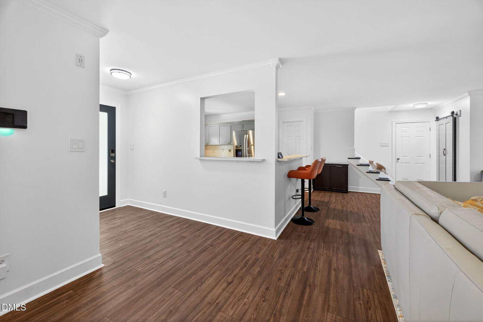 Modern open-concept living space with wood flooring, white walls, a kitchen window opening, bar stools at a counter, and a white sectional sofa near a barn door.