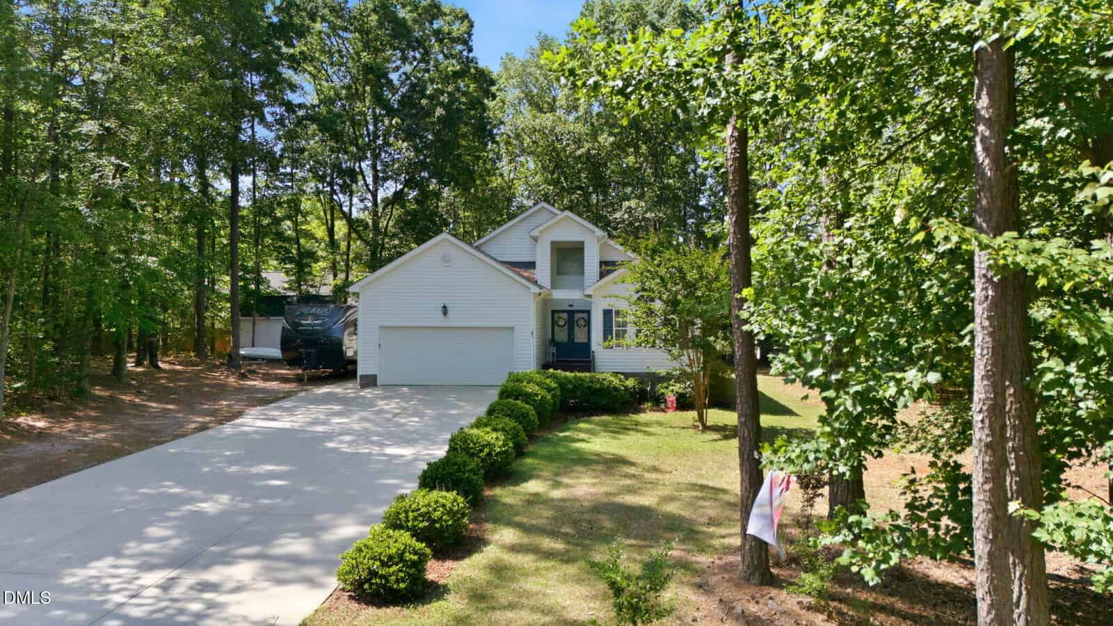 A white two-story house at 187 Buckaroo Drive in Louisburg features a blue front door, attached garage, and a long driveway. Surrounded by green trees and bushes on a sunny day, an RV is parked beside the house.