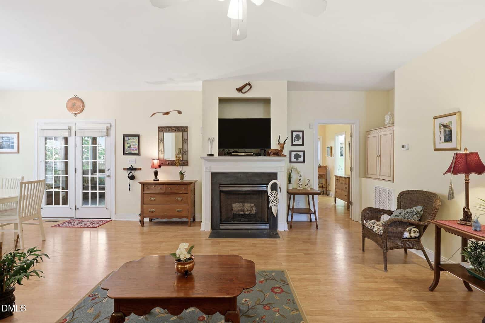 Bright living room with light wood floors, a central fireplace, TV, and artwork above; French doors, wicker chair, small tables, plants, and a dining area are also visible. The space feels open and inviting.