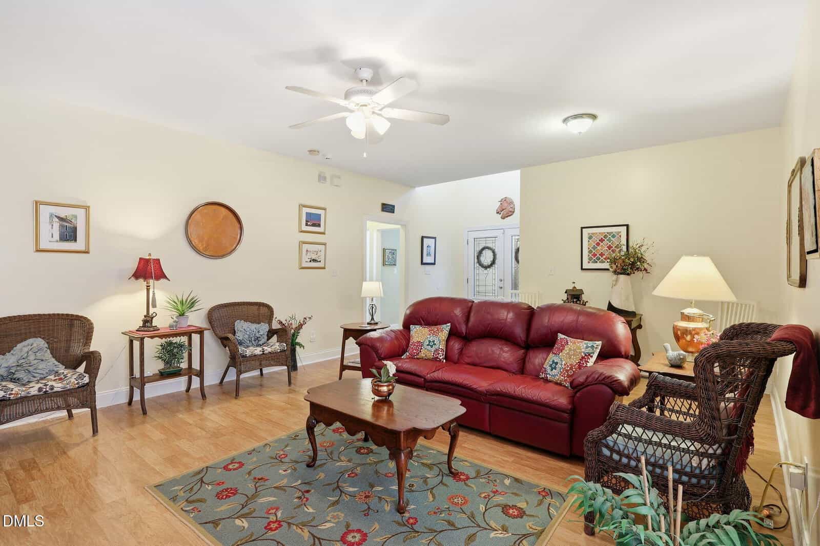 A cozy living room with a red leather sofa, wicker chairs, a wooden coffee table, floral rug, and several lamps. Light walls, wood flooring, framed art, and plants complete the inviting space.