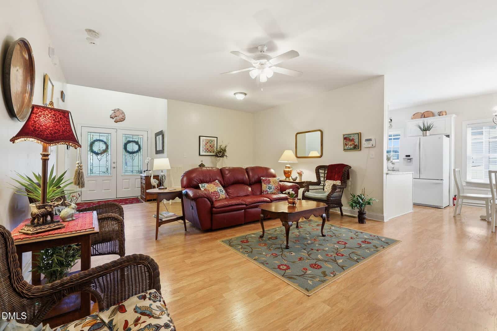 A cozy living room with a red leather sofa, wicker chairs, and a wooden coffee table on a floral rug. The space opens to a kitchen with white cabinets and appliances, and has light wood flooring throughout.