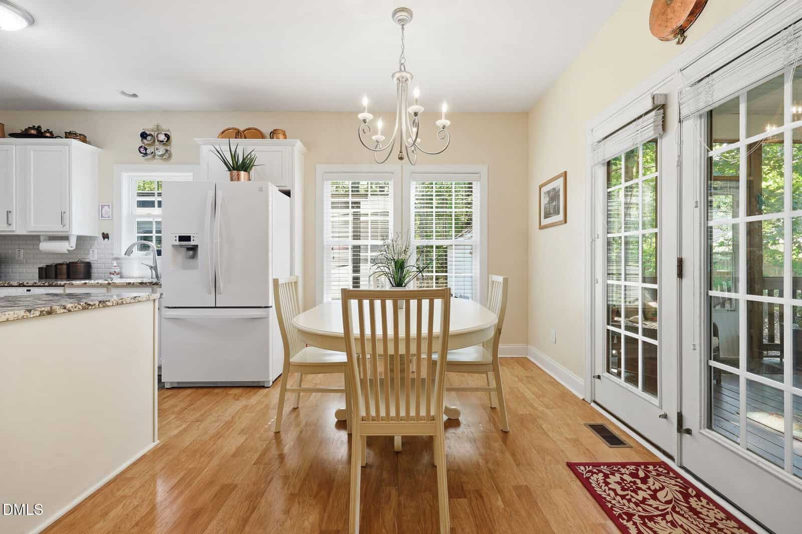 Bright kitchen with light wood floors, white cabinets, a white refrigerator, and a round dining table with four chairs. Large windows and glass doors let in natural light; a chandelier hangs above the table.
