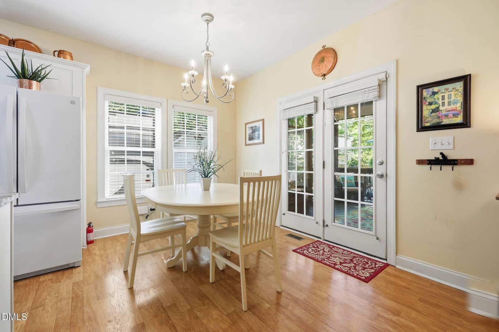 Bright kitchen with wood flooring, a round dining table with four white chairs, a white fridge, large windows, French doors leading outside, and a chandelier overhead. Plant and wall art add decorative touches.