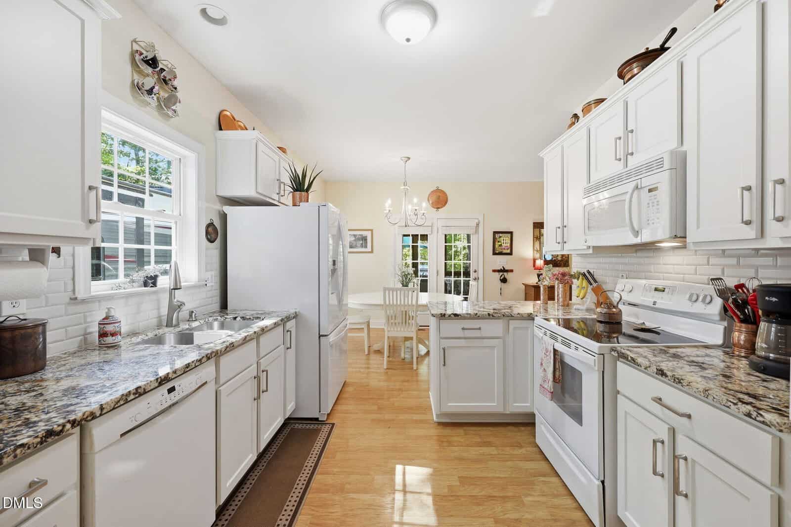 Bright kitchen with white cabinets, granite countertops, and stainless steel appliances. A dining area with a table and chairs is visible near glass doors that open to the outside. Sunlight streams through the windows.