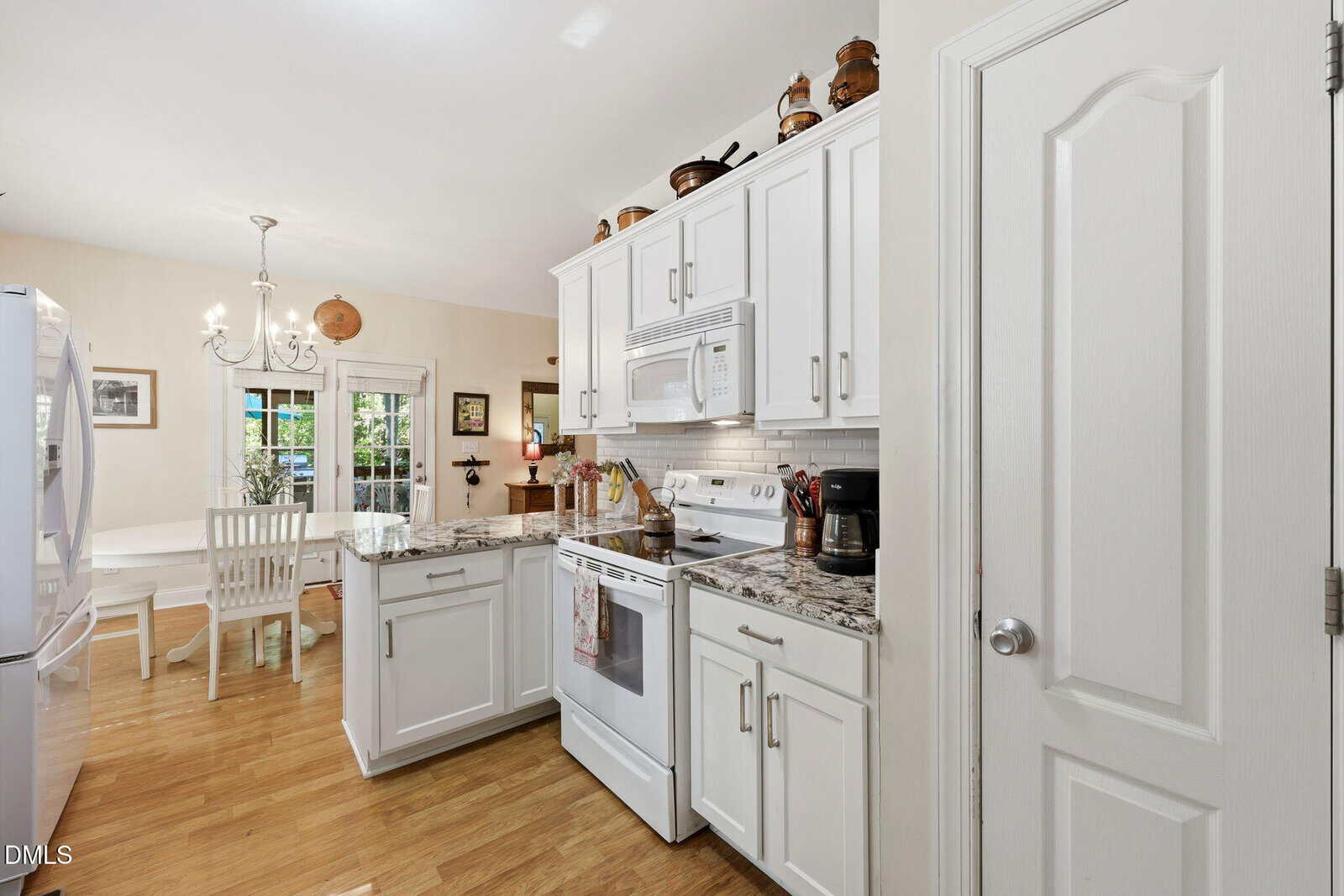 A bright kitchen with white cabinets, granite countertops, and stainless steel appliances. A dining table with chairs is near glass doors leading outside. Hardwood floors and decorative items are visible.