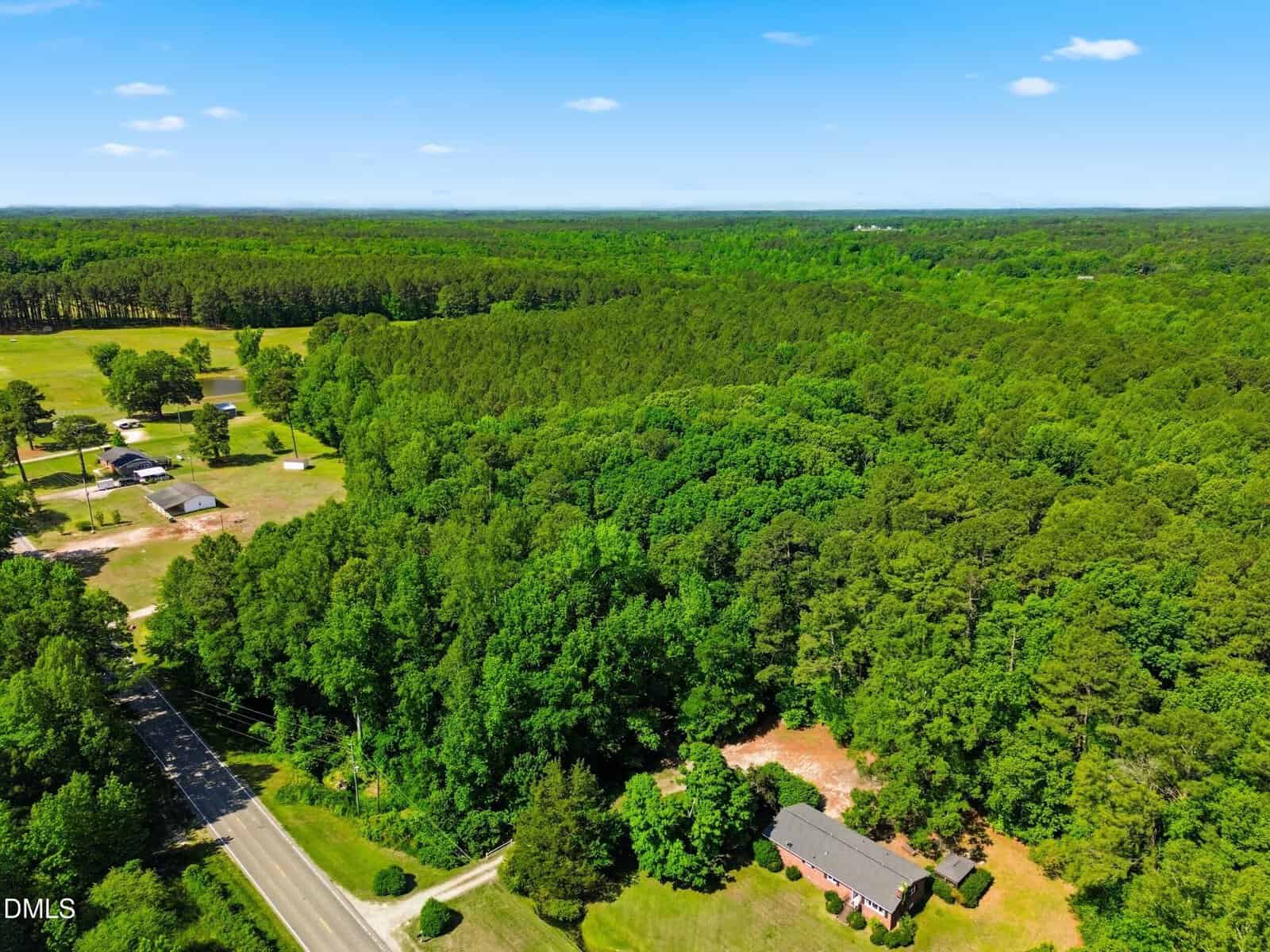 Aerial view of a house on 9.97 acres off S Hwy 39 in Zebulon, surrounded by dense green forest, with a road skirting the trees and a few nearby buildings under a clear blue sky.