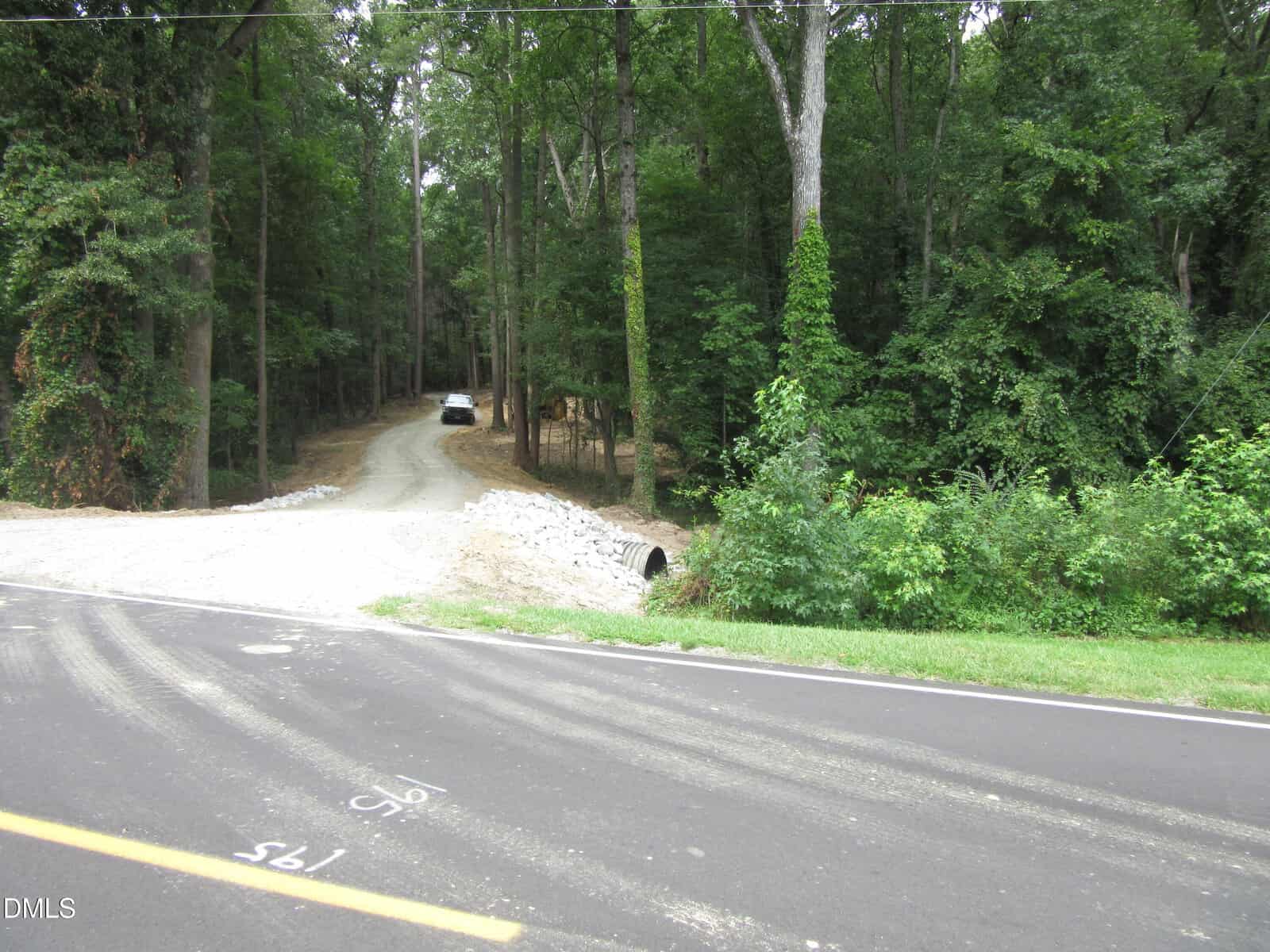 A white car is parked on a gravel driveway that curves into a dense, green forest. The driveway crosses a small culvert beside a paved road with numbers marked on its surface.