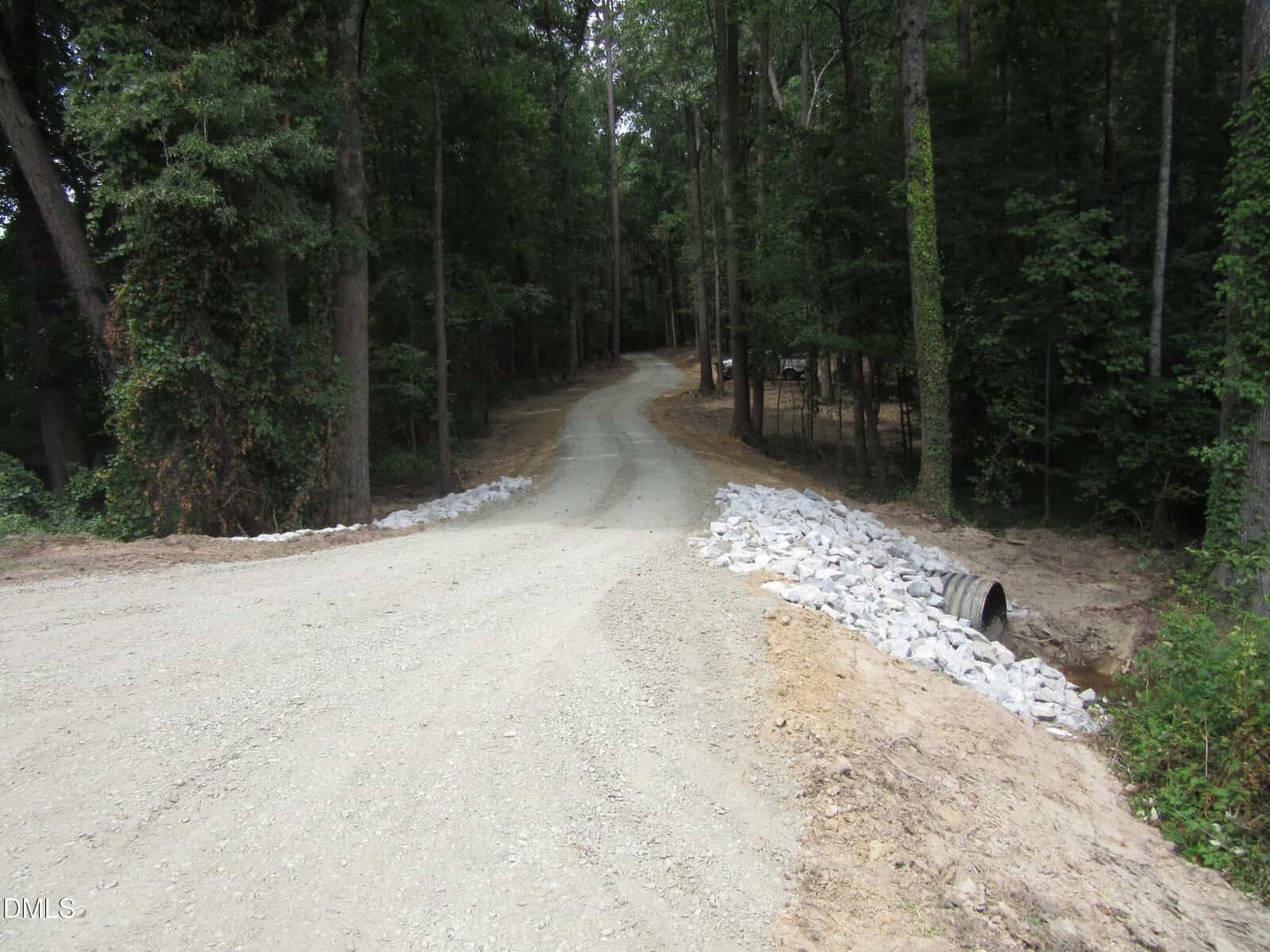 Gravel road surrounded by trees, curving to the right, with white rocks lining the edges and a drainage pipe visible on the right side.