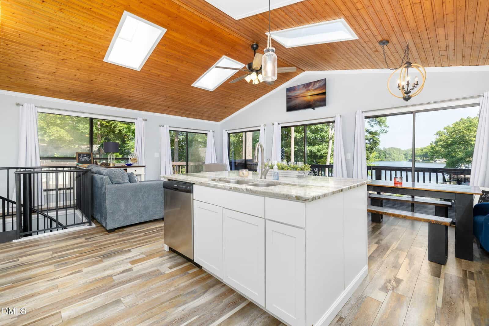 Modern open kitchen and living area with white cabinets, marble island, wood ceiling, skylights, large windows with lake views, and sliding doors opening to a deck. Dining table and gray sofa visible. Bright, airy atmosphere.