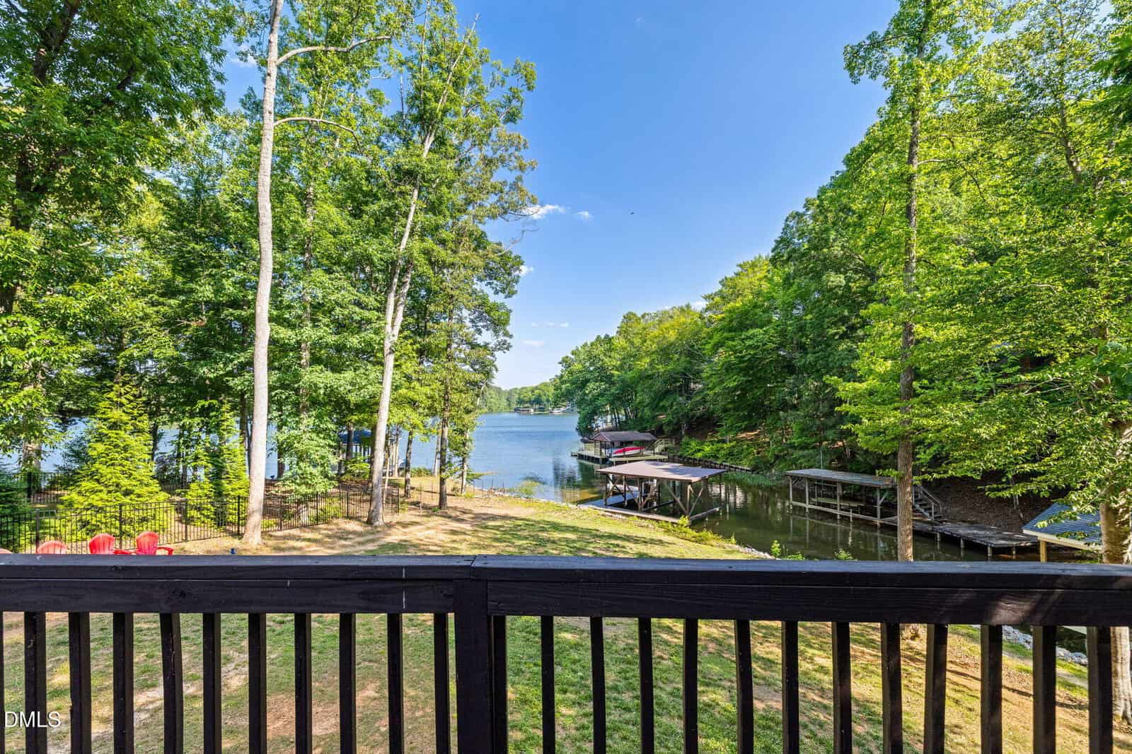 View from a wooden deck at 106 Bass Court, overlooking a peaceful lake surrounded by lush green trees in Macon, with docks and red Adirondack chairs visible by the shoreline on a sunny day.