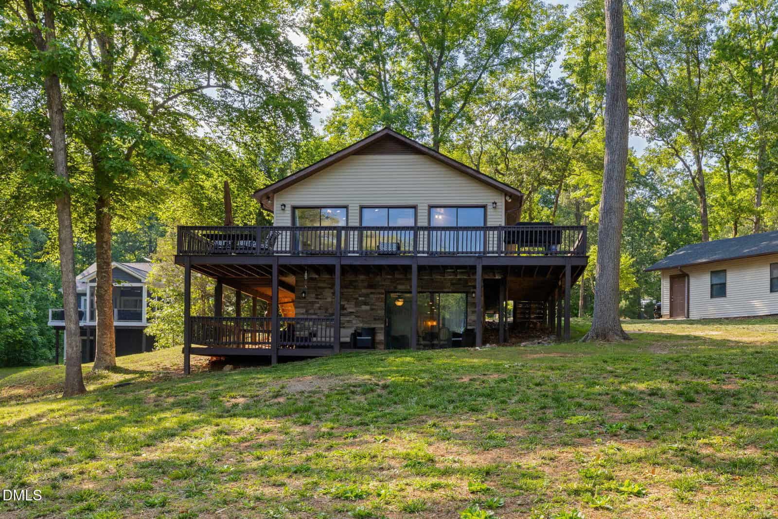 A two-story house with a large elevated deck and stone lower level sits among tall trees on a grassy lot, with another house visible to the right and one partially visible to the left.