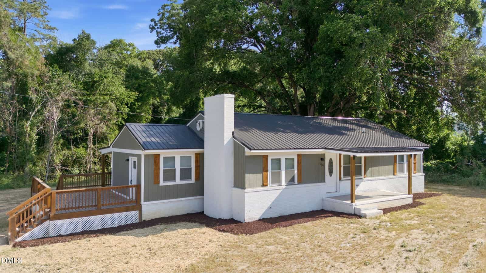 A small, single-story house with white brick and gray siding at 12835 NC 39 Highway N in Henderson, featuring a covered front porch, a side wooden deck, and surrounded by trees and grass under a clear blue sky.
