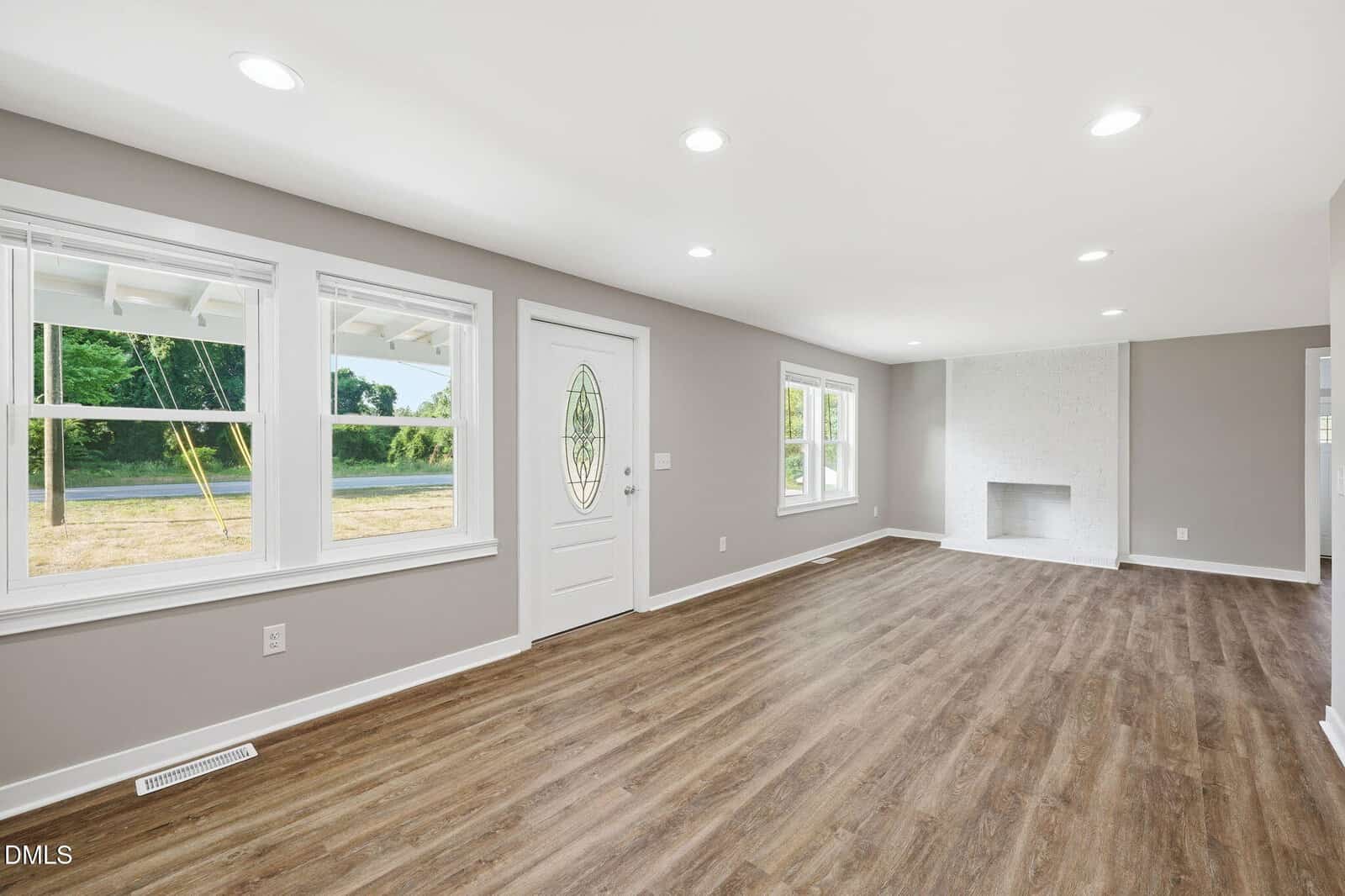 A bright, empty living room with light gray walls, three windows, a white door with an oval window, wood-style flooring, and a white brick fireplace. Recessed ceiling lights are evenly spaced throughout the space.