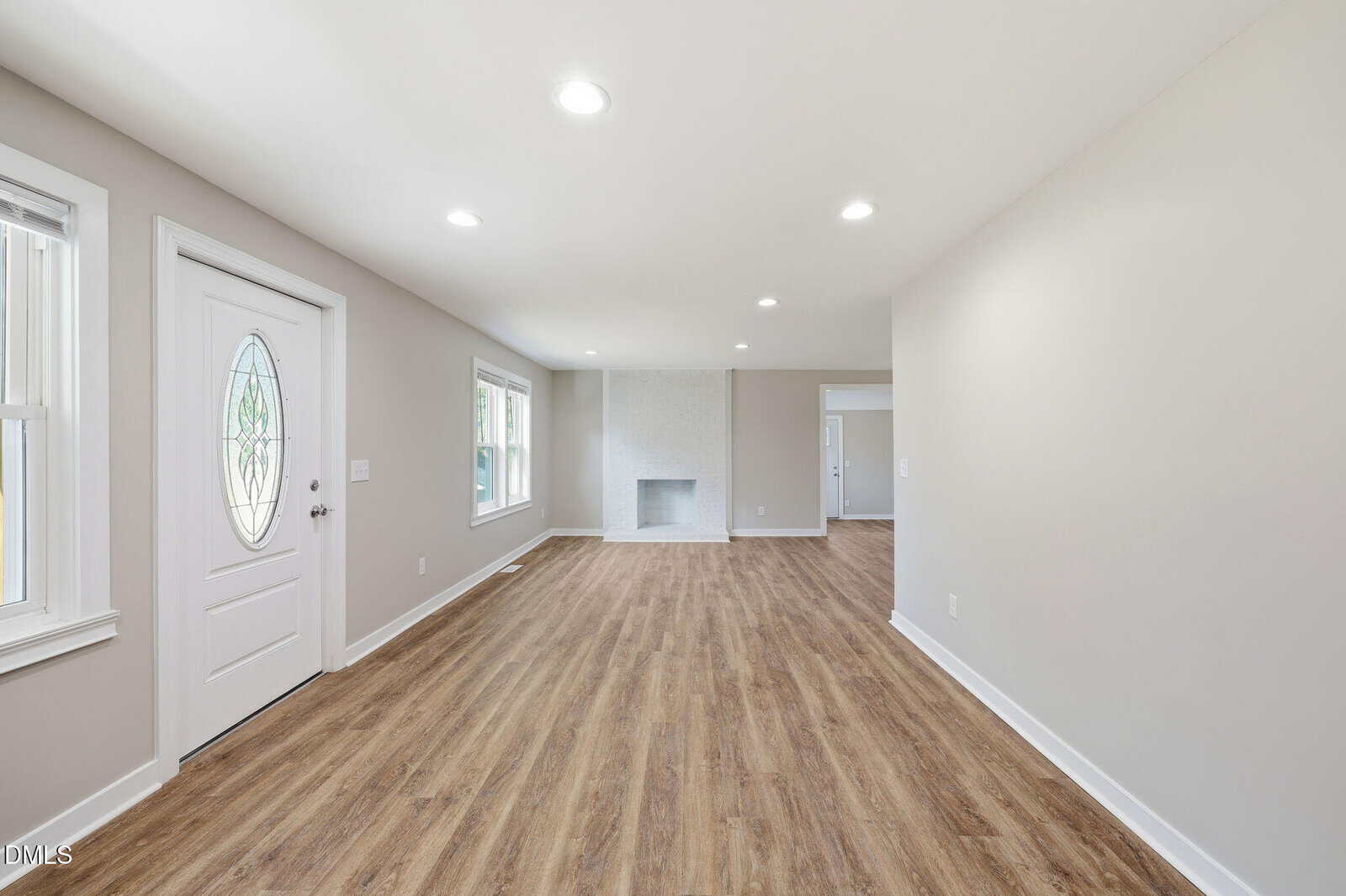 A spacious, empty room with light-colored walls, wood-look flooring, recessed ceiling lights, a white front door with a window, and large windows allowing natural light. A fireplace is visible at the far end of the room.