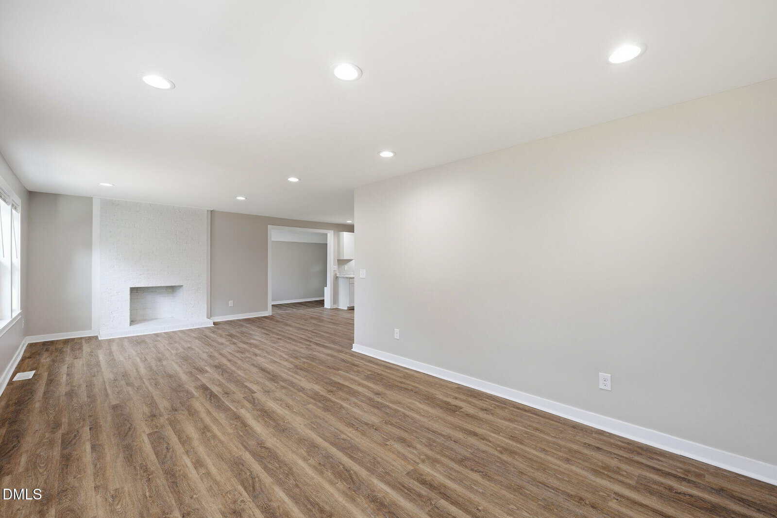 Open living space with light wood flooring, neutral walls, recessed lighting, and a white brick fireplace. The area connects to another room, with large windows along one wall allowing natural light to enter.