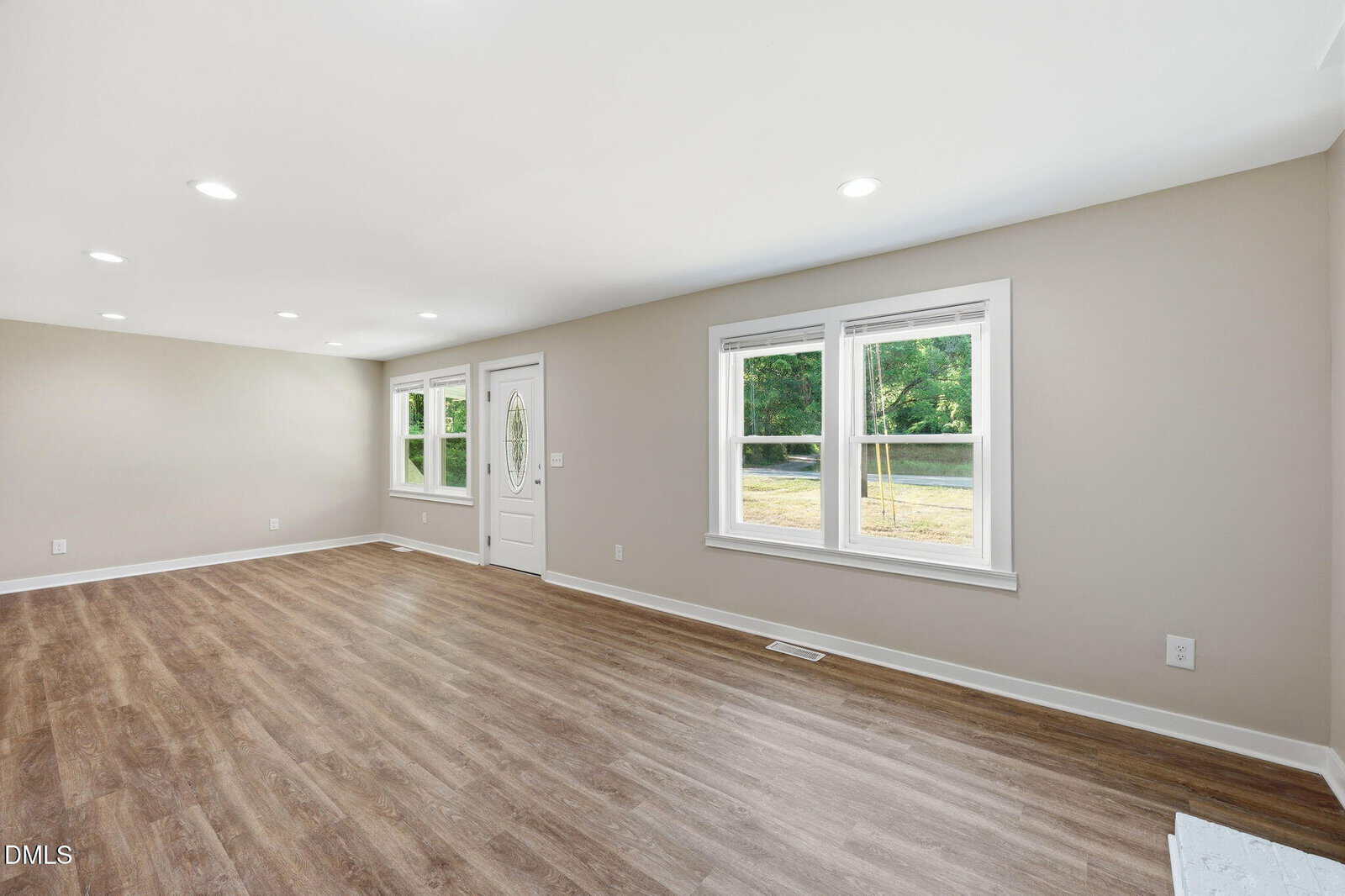 Bright, empty living room with light wood floors, beige walls, white trim, recessed lighting, and two windows letting in natural light. White door with oval glass panel is near the center.