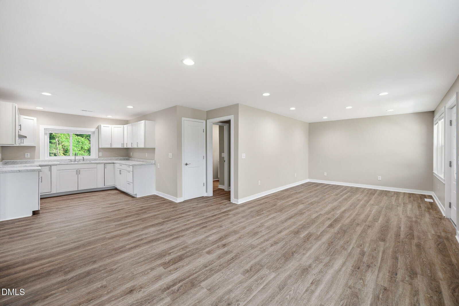 Spacious open-plan room with wood floors, white walls, recessed lights, and a modern white kitchen with large windows and ample cabinetry.