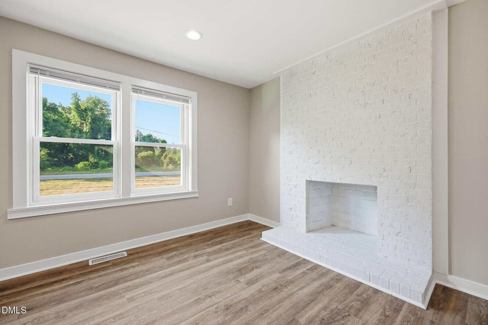 A bright room with light wood flooring, beige walls, a large double window, and a white painted brick fireplace with a raised hearth.