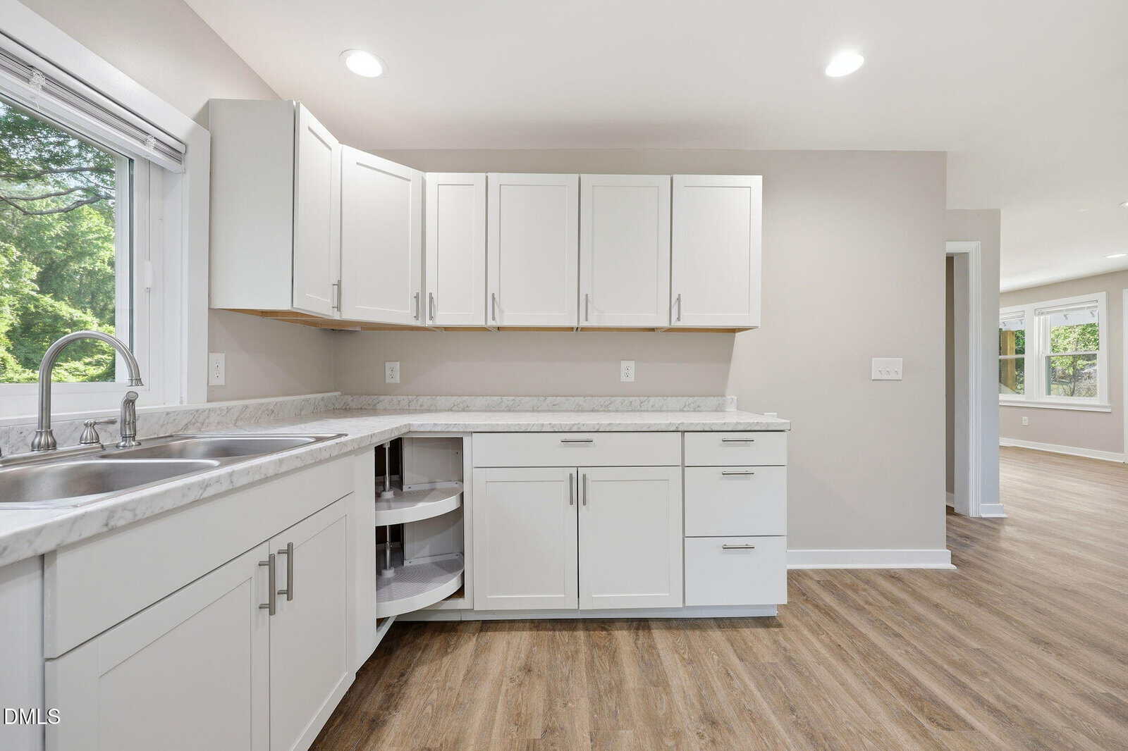 Modern kitchen with white cabinets, marble countertops, double sink, corner cabinet with a rotating shelf open, and light wood flooring. Large window brings in natural light and greenery from outside.