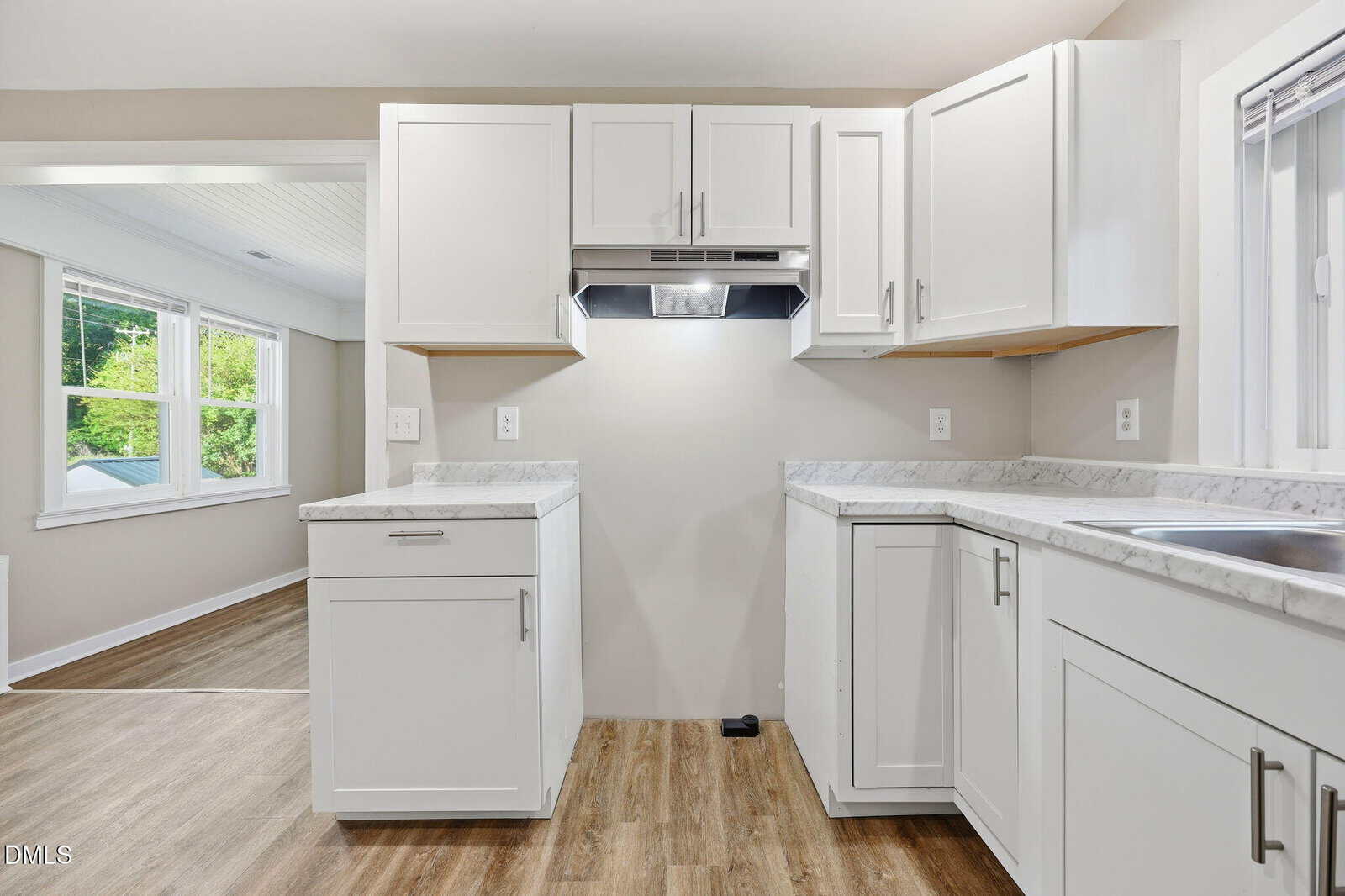Modern kitchen with white cabinets, marble countertops, stainless steel range hood, wood flooring, and a window showing a bright, green outdoor view. The kitchen opens into a sunlit adjacent room.