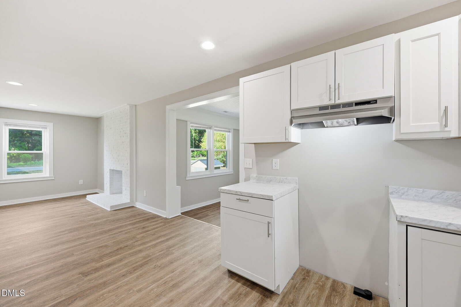 Modern, open-concept living space with light wood flooring, white walls, and white kitchen cabinets; large windows provide natural light and a view of greenery outside. Fireplace visible in the living area.