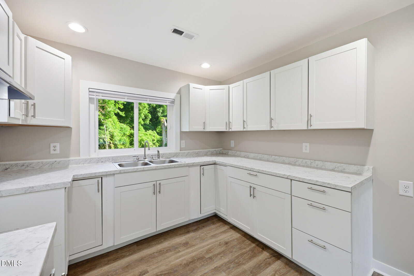 A bright kitchen with white cabinets, marble countertops, stainless steel double sink under a window, and wood-style flooring. The window offers a view of green trees outside.