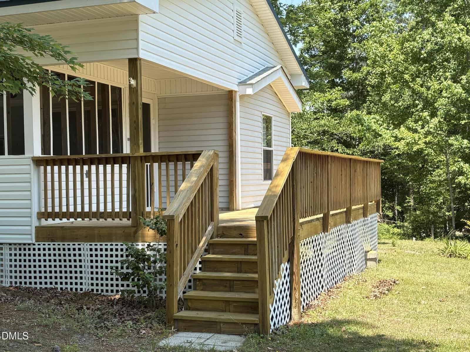 Wooden steps and a ramp with railings lead to the entrance of a white house with a porch. The house is surrounded by green trees and grass, with lattice skirting around the porch foundation.