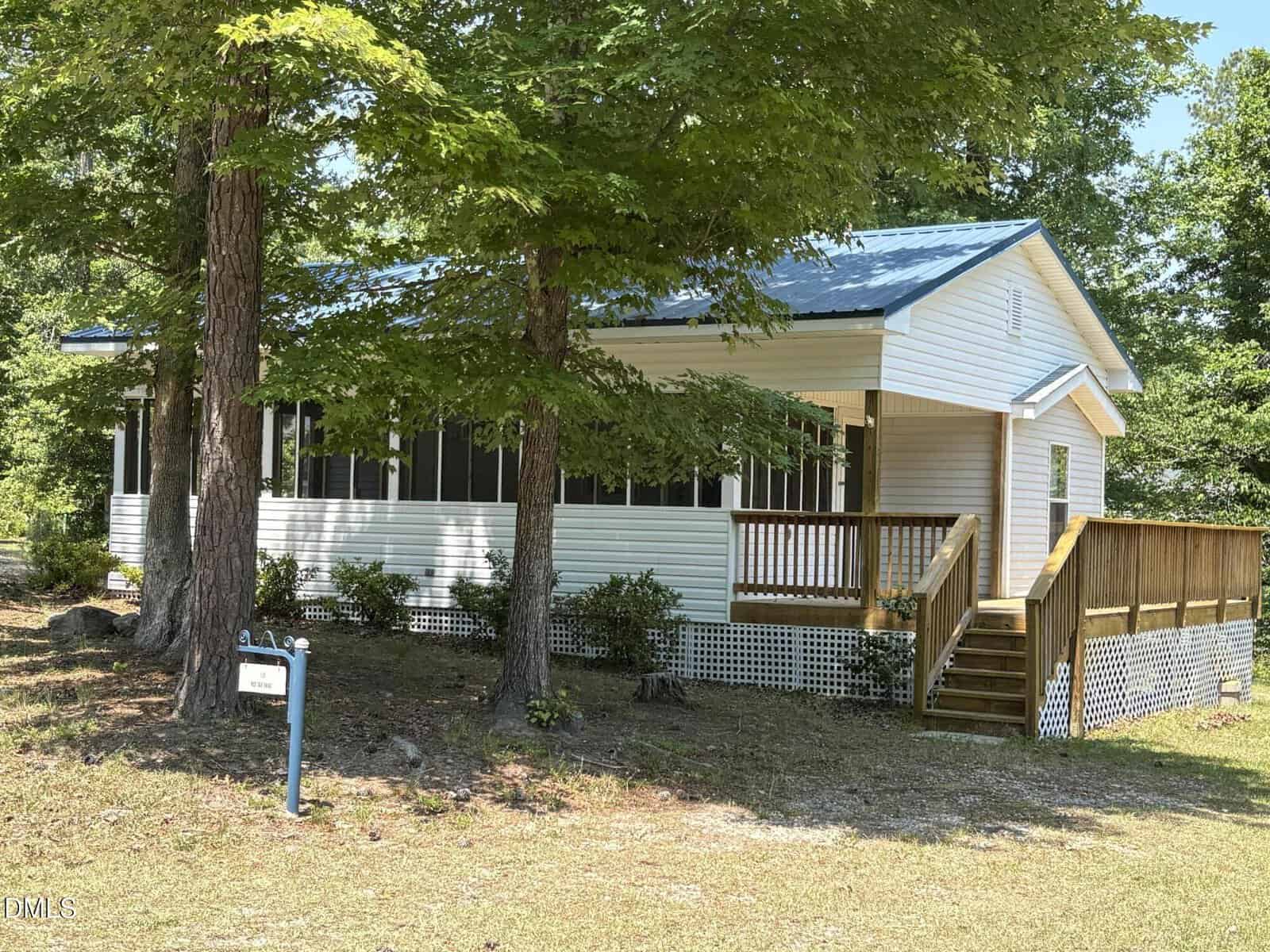 A small white house with a blue metal roof and screened-in front porch sits at 115 Red Oak Drive, Louisburg. Surrounded by trees and greenery, wooden steps lead to the porch, and a signpost is visible in the yard.