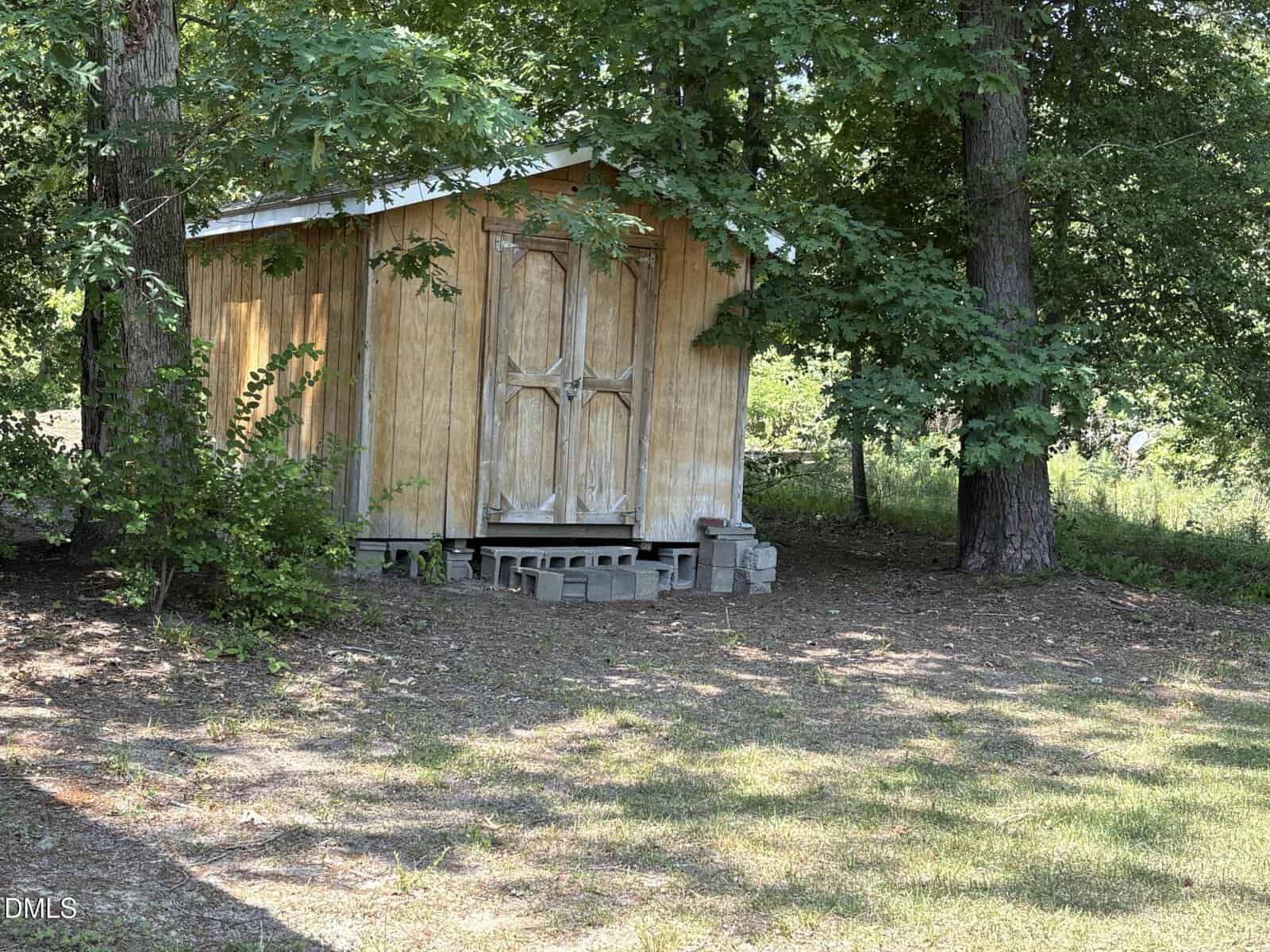 A small wooden shed with double doors stands on concrete blocks under the shade of trees, surrounded by grass and scattered patches of sunlight.