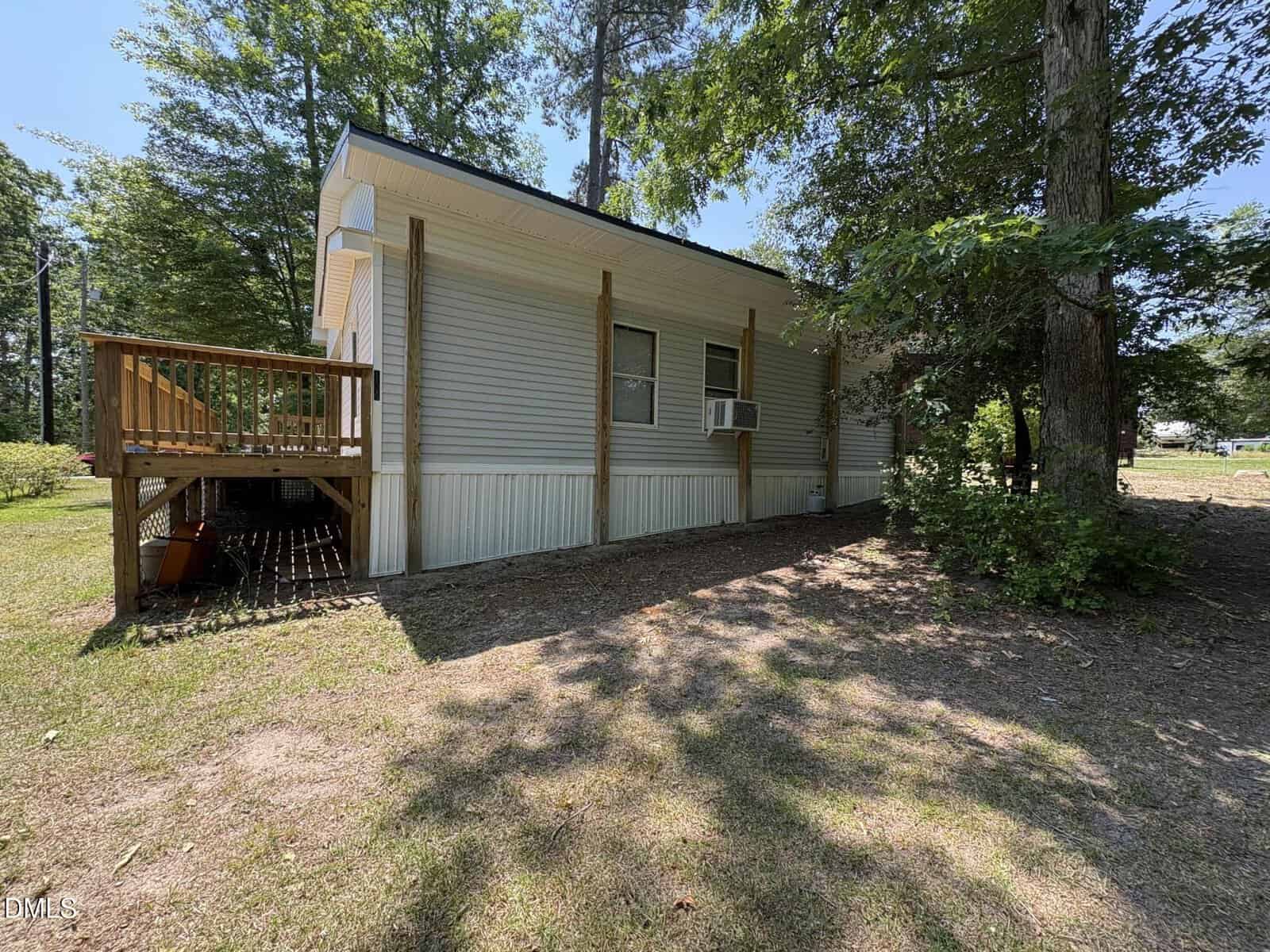A small, light-gray house with a wooden deck and an air conditioning unit in the window, surrounded by trees and grass on a sunny day. Shadows from trees are cast on the ground.