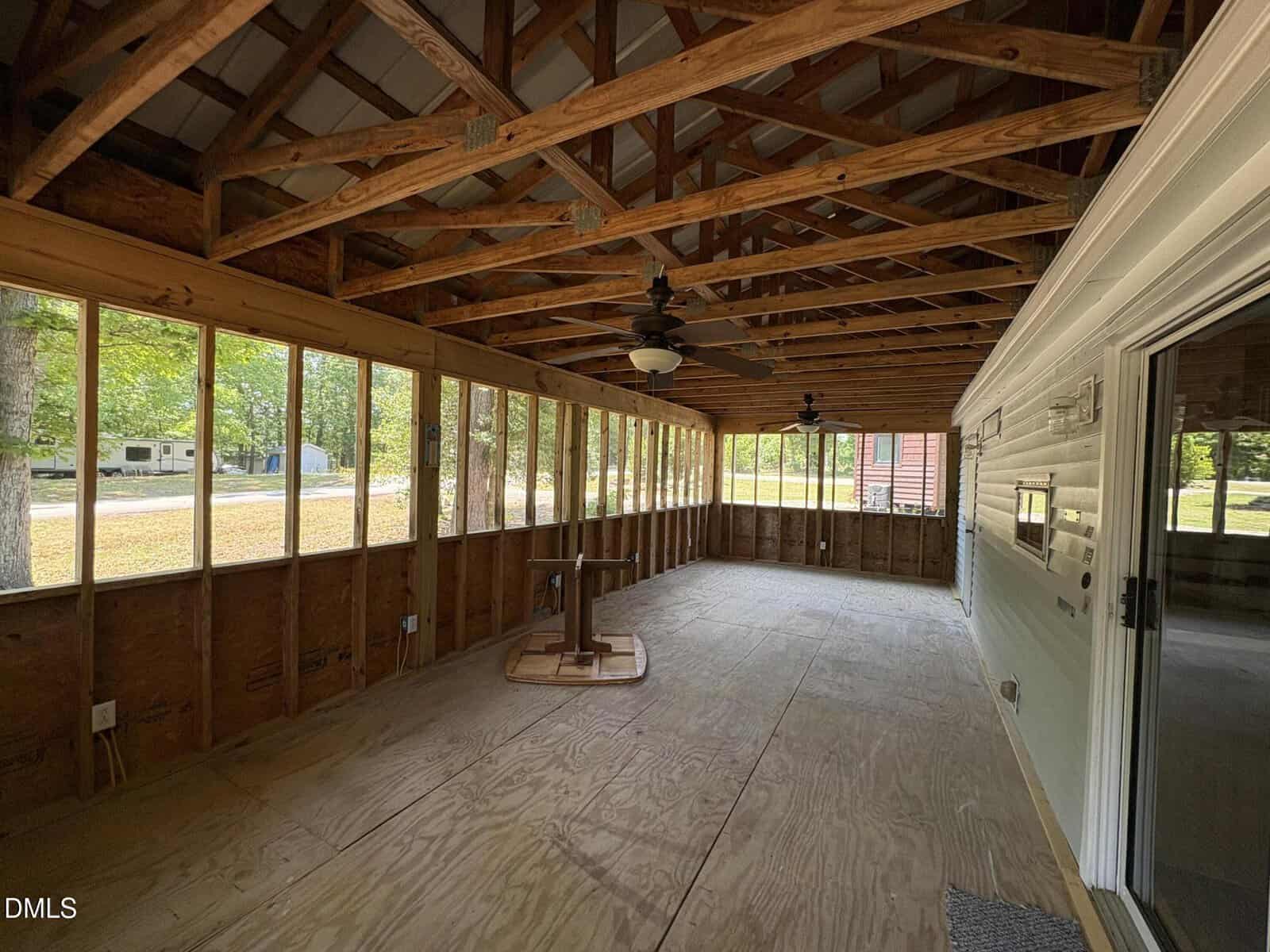 A spacious screened-in porch with unfinished wooden floors and exposed ceiling beams, featuring ceiling fans, overlooking a yard with trees. A small table is upside down on the floor.