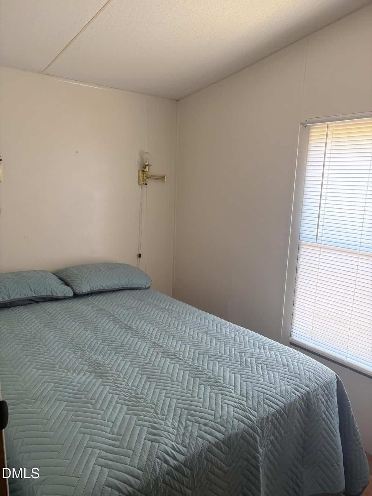 A small bedroom with a neatly made bed covered in a blue quilt, two pillows, a beige wall, a wall-mounted lamp, and sunlight filtering through closed blinds on the window.