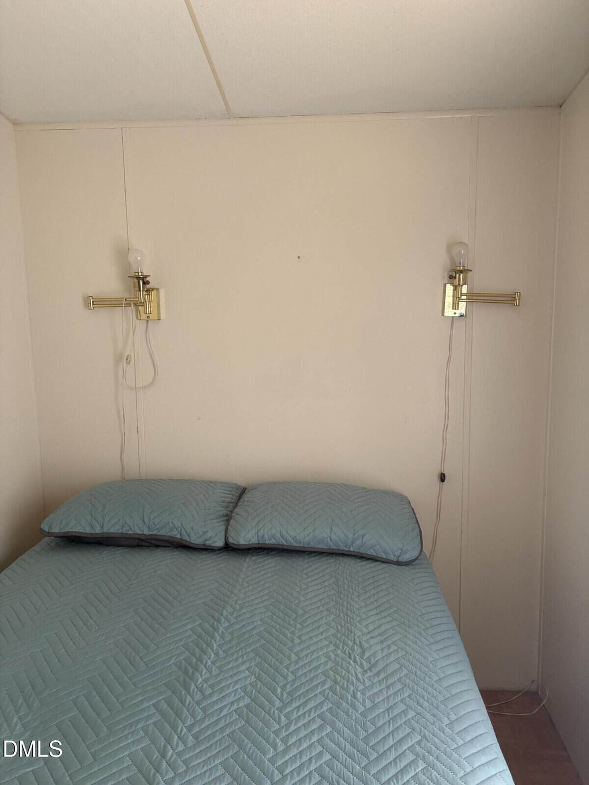 A bed with a light blue quilt and two pillows is centered against a plain cream wall. Two wall-mounted brass lamps are above the bed, one on each side, with exposed bulbs.