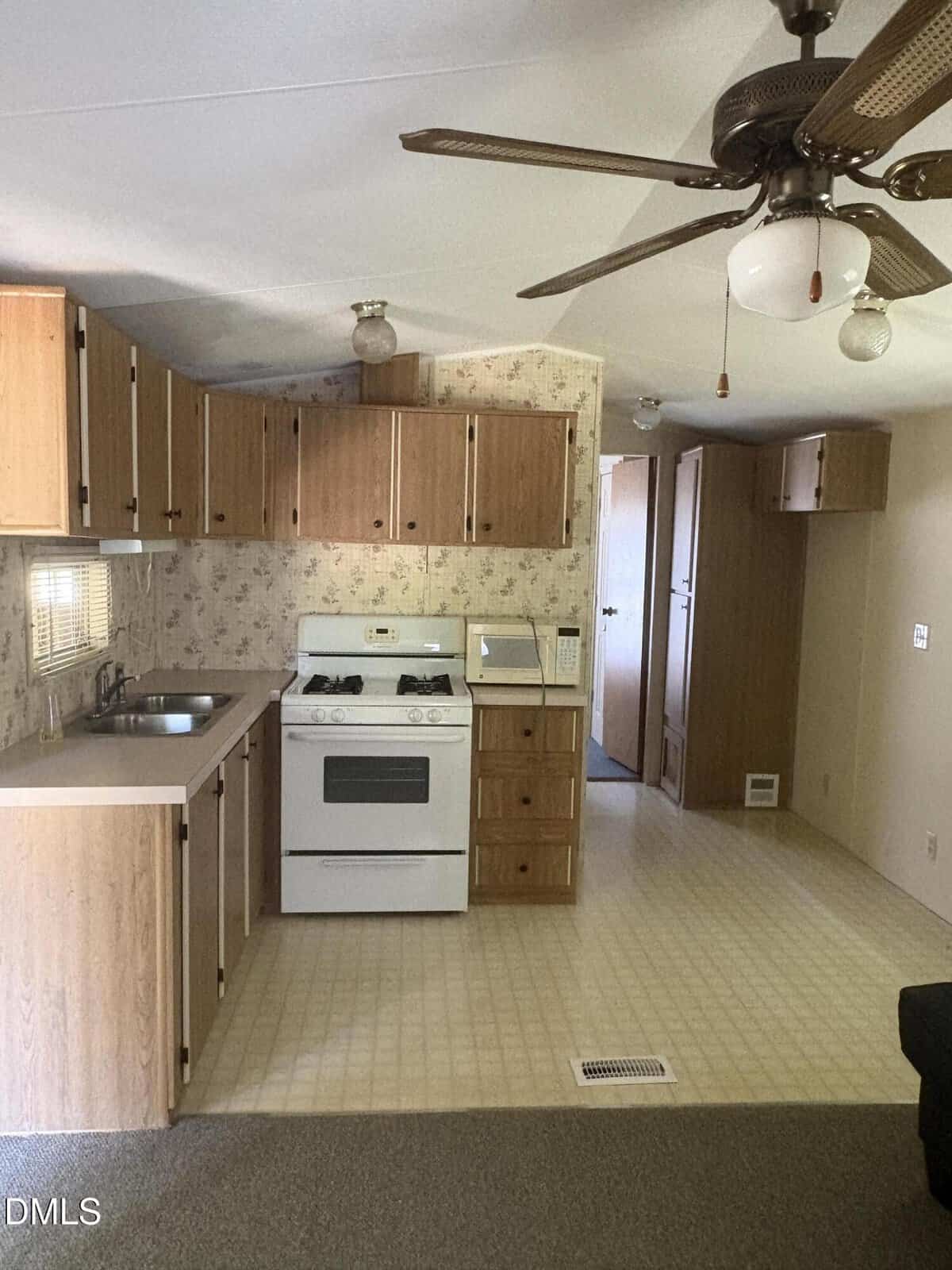 Small kitchen with wood cabinets, gas stove, sink, and microwave. Ceiling fan with a light is visible. Brown carpet meets vinyl flooring; wallpaper with a floral pattern lines the wall. Doorway leads to another room.