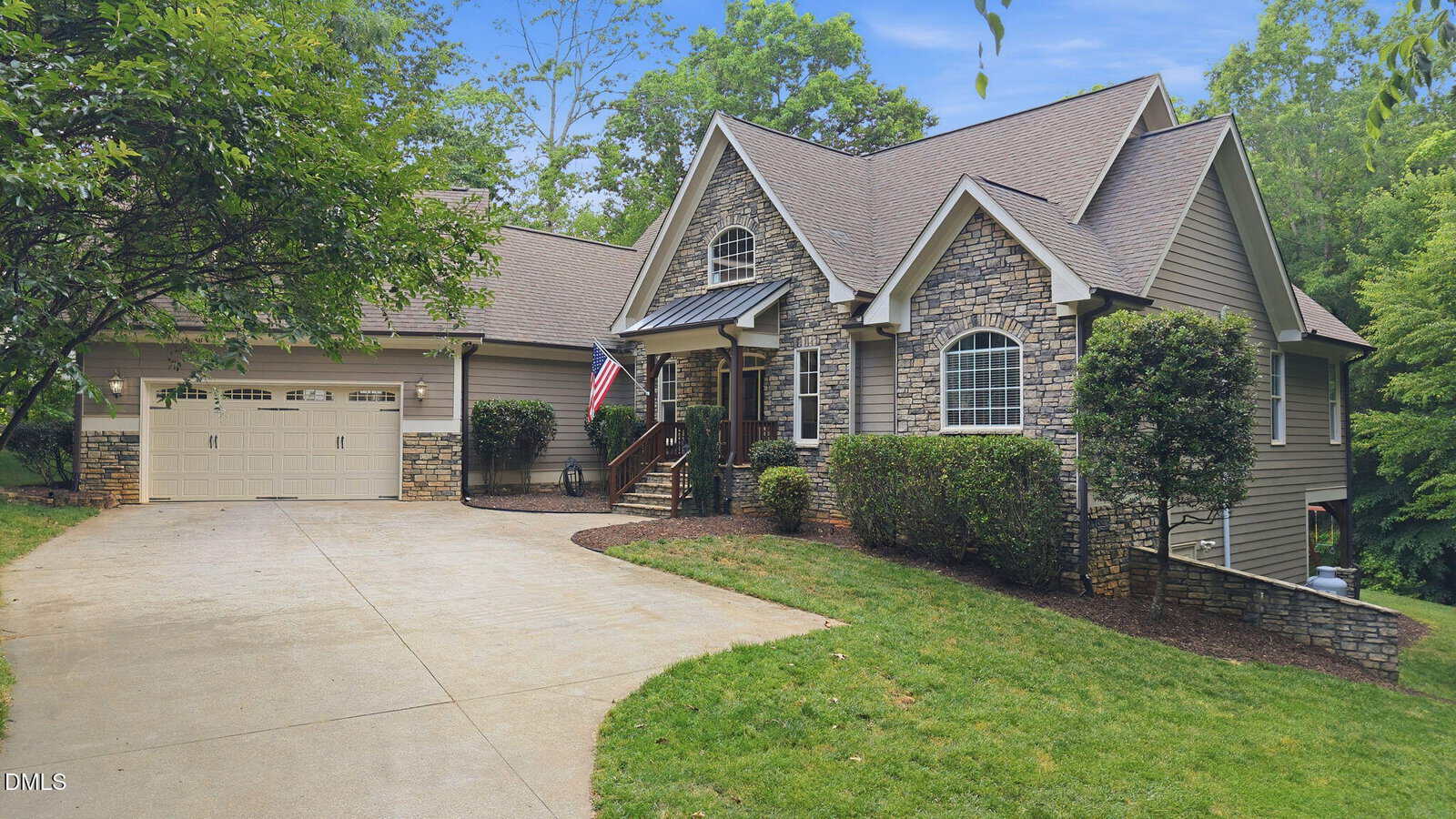 A suburban house in Youngsville at 1001 Browning Place, featuring a stone and siding exterior, gabled roof, attached two-car garage, American flag by the front porch, lush trees and shrubs, and a curved concrete driveway.