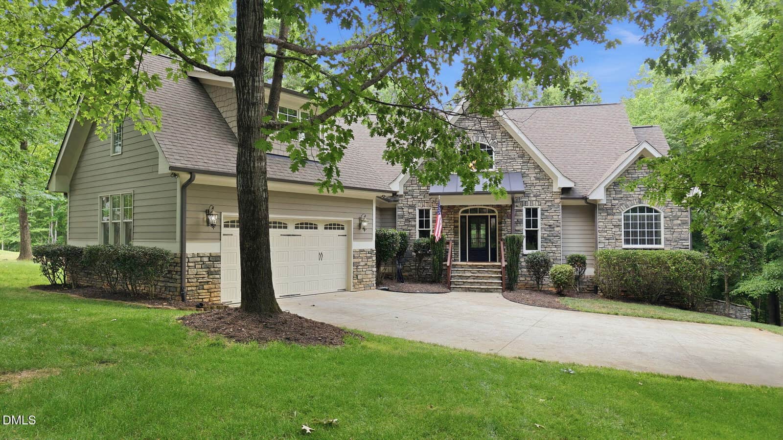 A two-story suburban house with stone and siding exterior, an attached two-car garage, and a manicured front lawn surrounded by trees and shrubs on a sunny day.