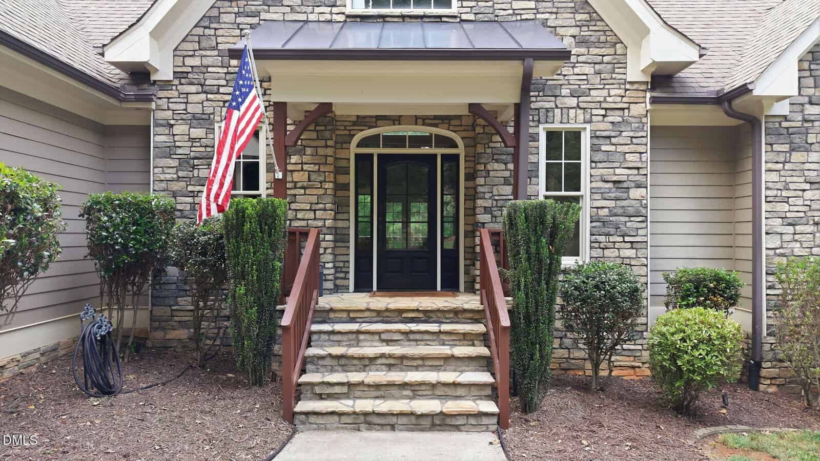 Front entrance of a house with stone and siding exterior, a black front door with windows, steps leading to a small porch, an American flag, and trimmed bushes on either side of the steps.