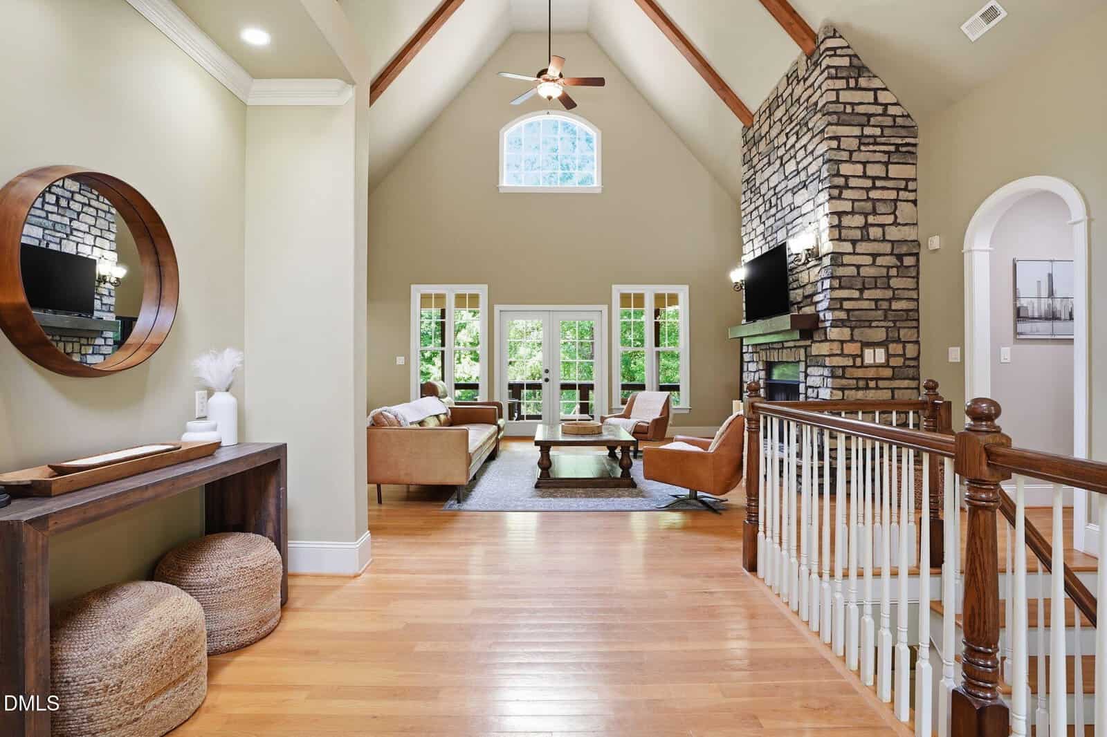 Open-concept living room with vaulted ceiling, exposed beams, stone fireplace, tan walls, hardwood floors, large windows, brown sofas, a round mirror above a console table, and a staircase with white spindles.