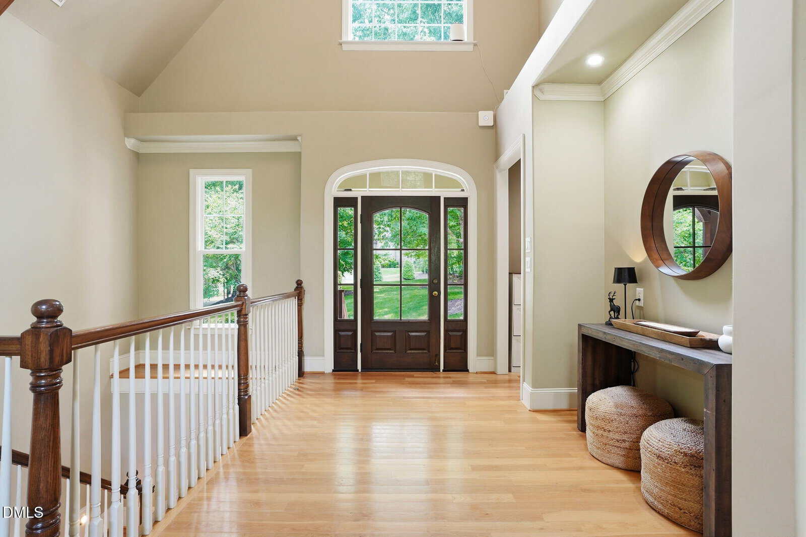 A spacious, bright hallway with polished wood floors, a dark wood front door with glass panels, a staircase with white spindles, a console table with a round mirror, and two woven ottomans underneath.