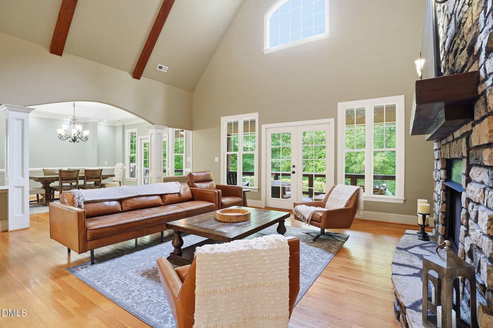 Spacious living room with tall ceiling, large stone fireplace, brown leather furniture, wood floors, area rug, and wide windows letting in natural light; dining area visible in the background through an archway.