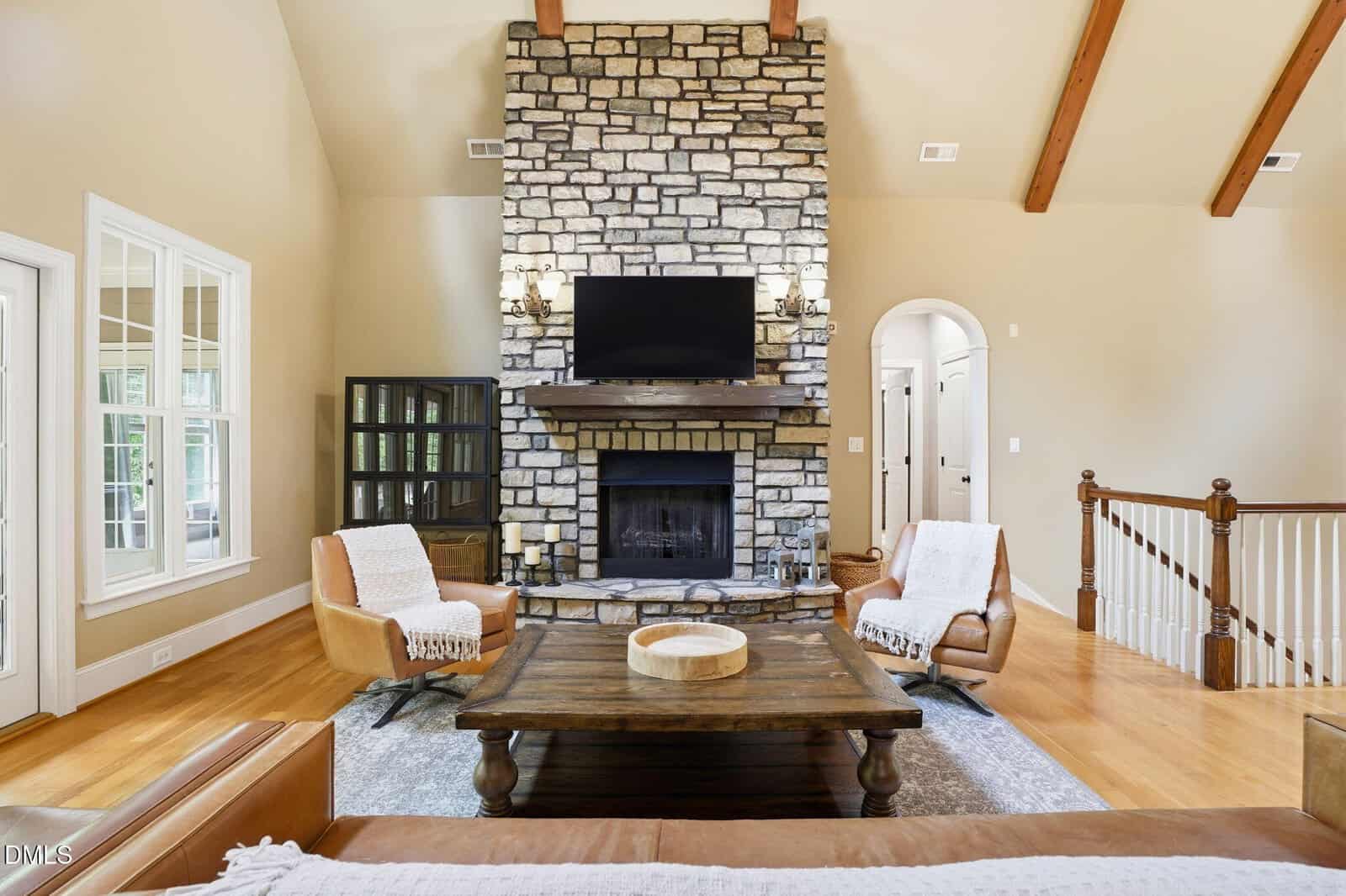 Living room with a stone fireplace, wall-mounted TV, two tan chairs, wooden coffee table, cabinet, and beige walls. Ceiling features wooden beams, and stairs with a wooden railing are visible on the right.