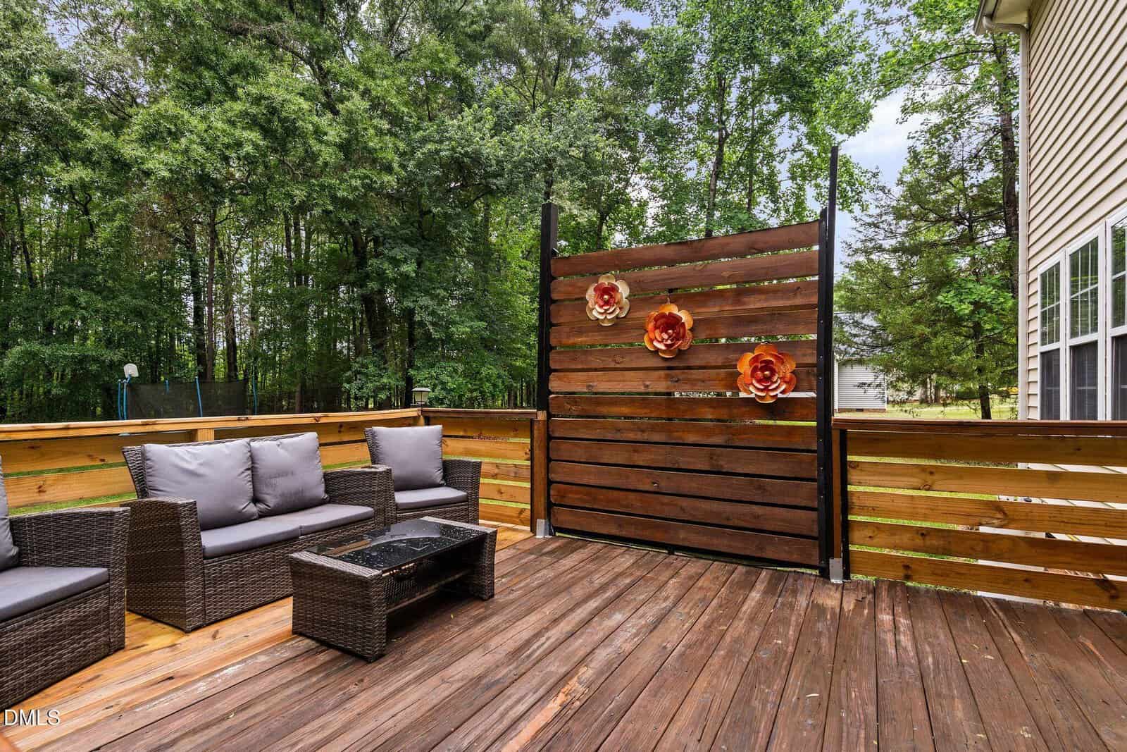 A wooden patio deck with outdoor wicker furniture, a coffee table, and a privacy screen decorated with three large flower ornaments, surrounded by trees and next to a house.